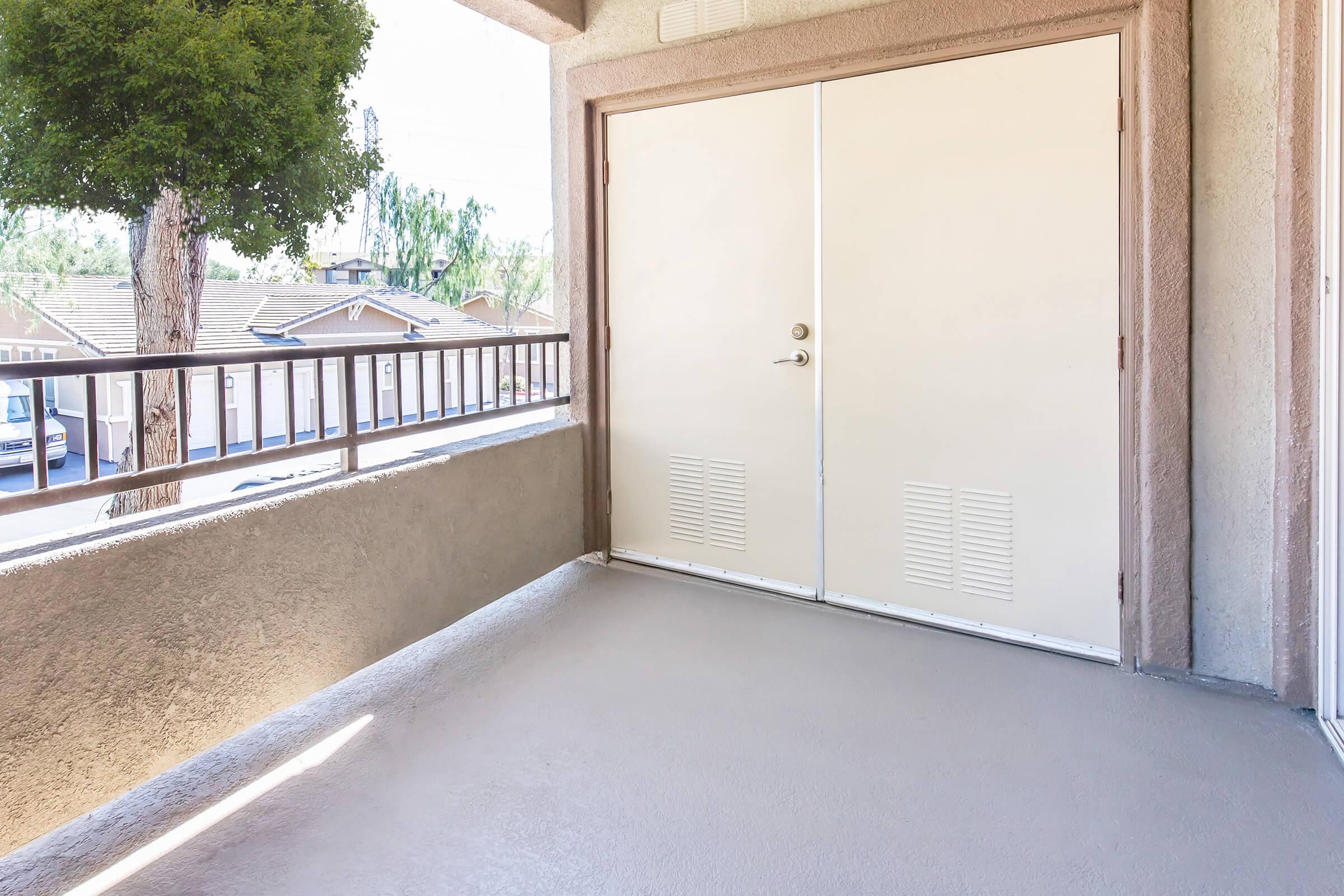 A view of a balcony with a light-colored concrete floor and a closed double door leading inside. The balcony has a railing and a tree is visible in the background, along with houses in the distance. Natural light streams in, creating a bright atmosphere.