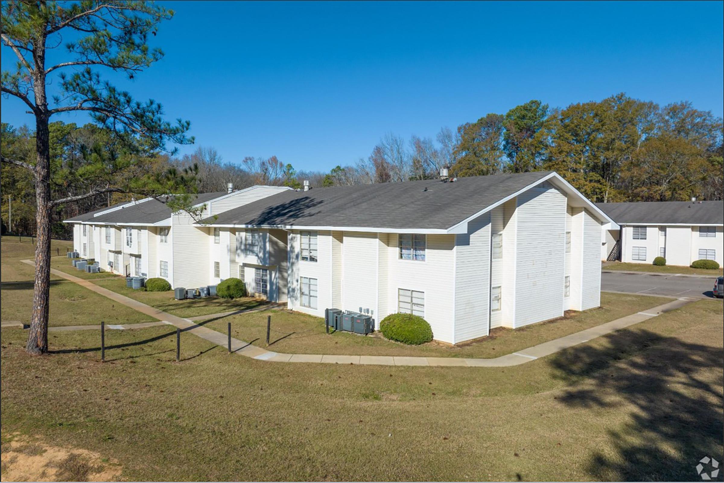 A row of white multi-family apartment buildings surrounded by grass and trees, with a clear blue sky overhead. The buildings feature windows and air conditioning units, set in a peaceful suburban environment. Light traffic can be seen on the nearby road.