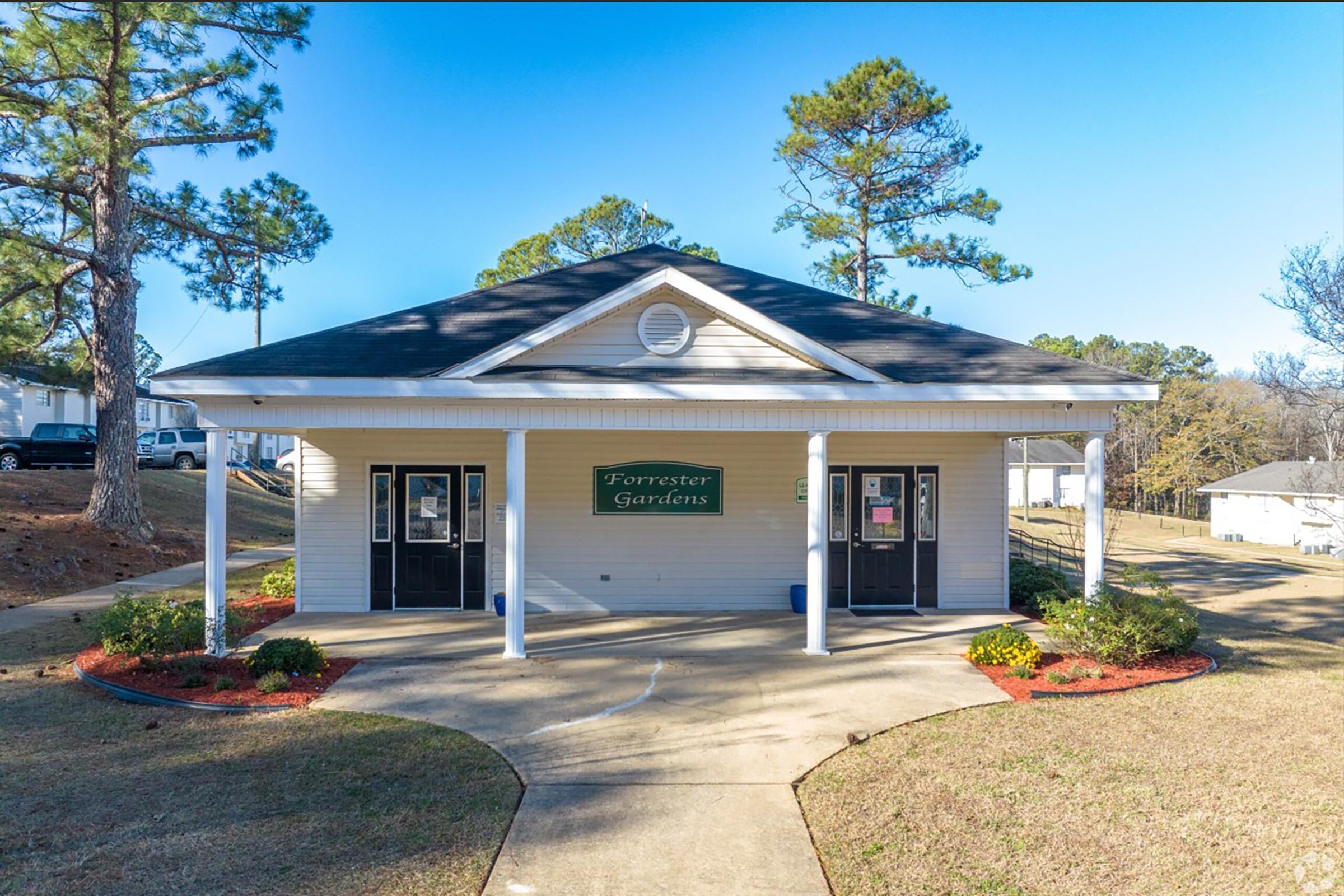 A single-story building with a pitched roof, featuring a sign that reads "Forrester Gardens." The entrance has two doors, and the surrounding area includes a tidy lawn with flower beds. Pine trees and other greenery are visible in the background against a clear blue sky.