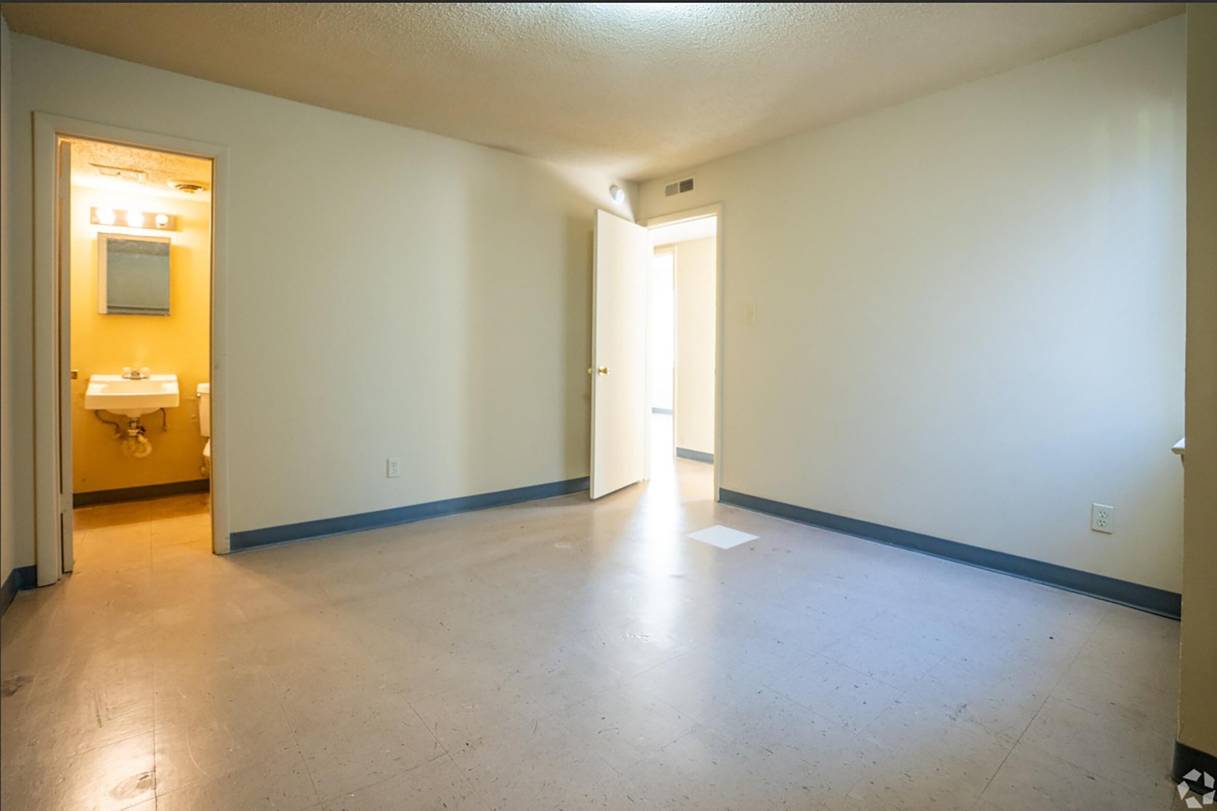A vacant room with beige walls and a gray floor. A doorway leads to a bathroom on the left, featuring yellow walls and a mirror. Another open doorway on the right leads to a hallway. The room is well-lit with natural light and appears spacious, providing a blank canvas for decoration.