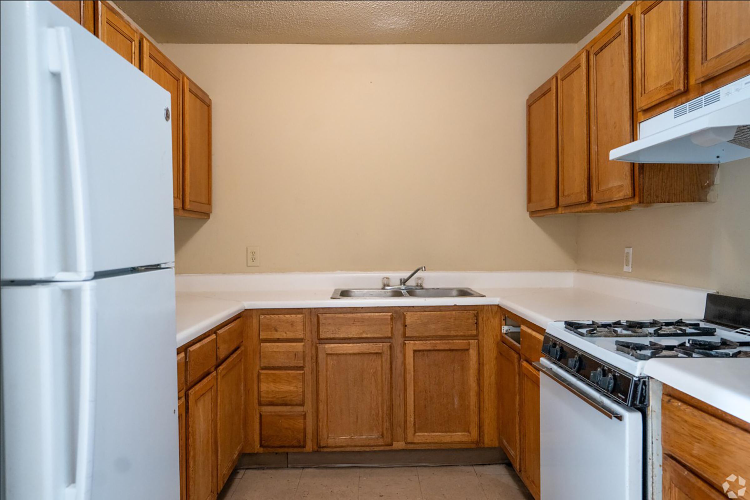 A kitchen featuring wooden cabinets, a white refrigerator, a stainless steel sink, and a stove with an oven. The walls are painted light beige, and the countertops are white. The kitchen is well-lit with a practical layout.