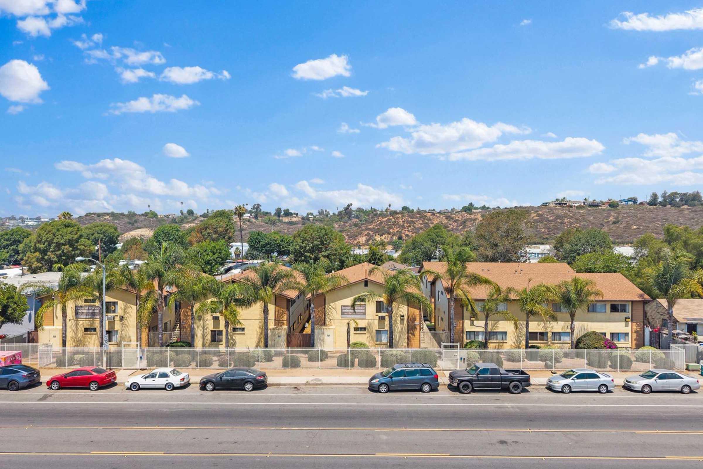 A view of a modest apartment complex featuring palm trees in front, set against a backdrop of rolling hills under a blue sky with scattered clouds. Several parked cars line the street in front of the complex, which has a warm color palette and a relaxed, suburban atmosphere.