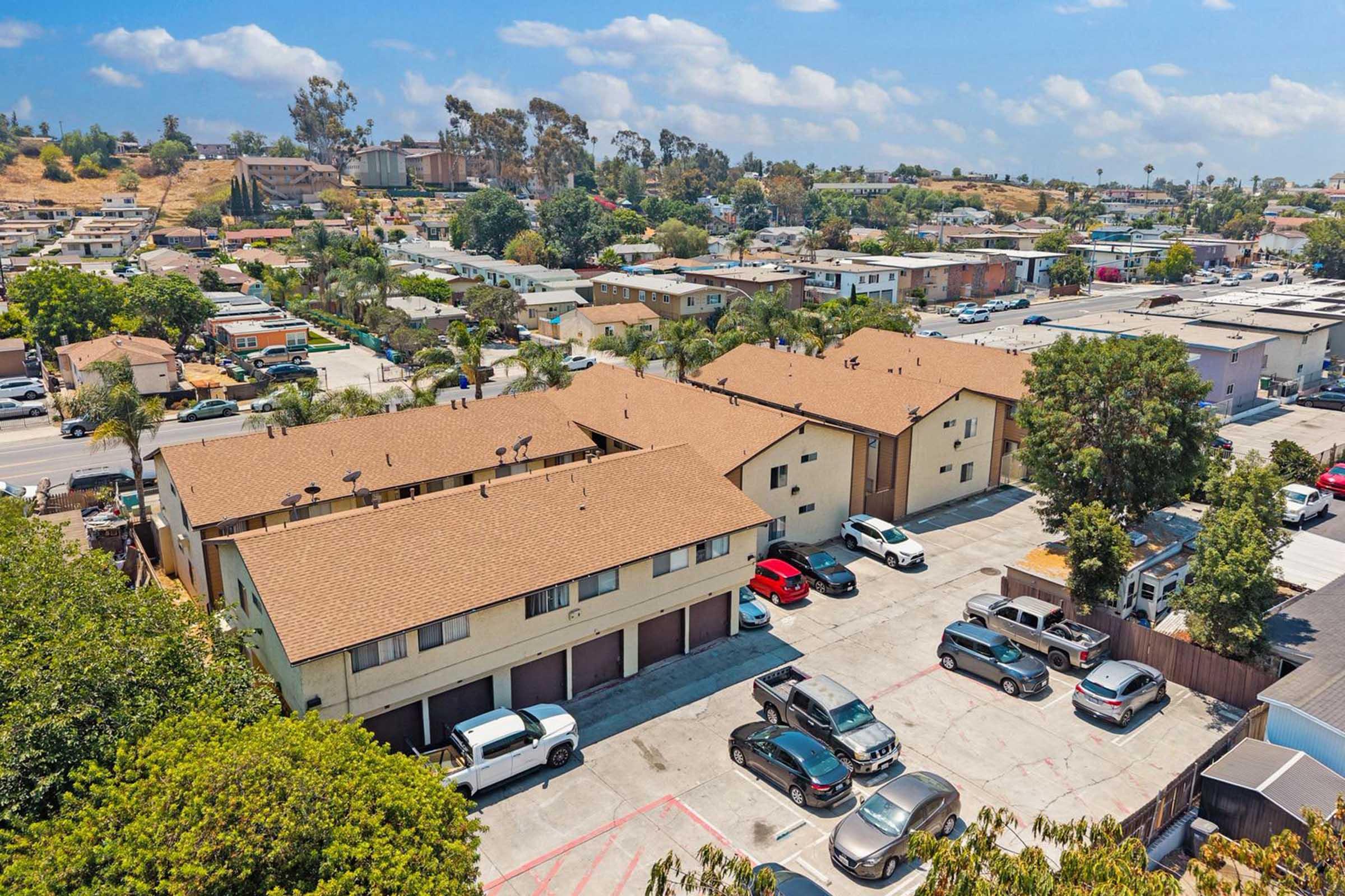 Aerial view of a residential area featuring multiple brown-roofed buildings, surrounded by greenery and parked cars in a lot. The background shows a mixture of houses and commercial buildings under a blue sky with scattered clouds.