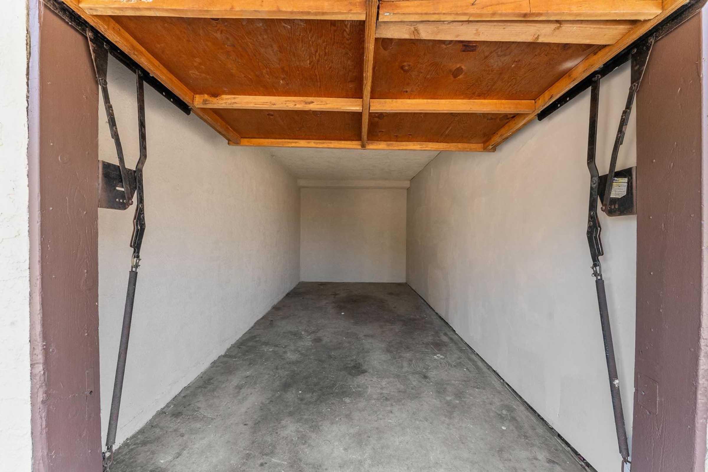 An empty garage with a concrete floor and whitewashed walls. The overhead door is partially open, revealing a wooden ceiling structure. The space is uncluttered and ready for storage or parking.