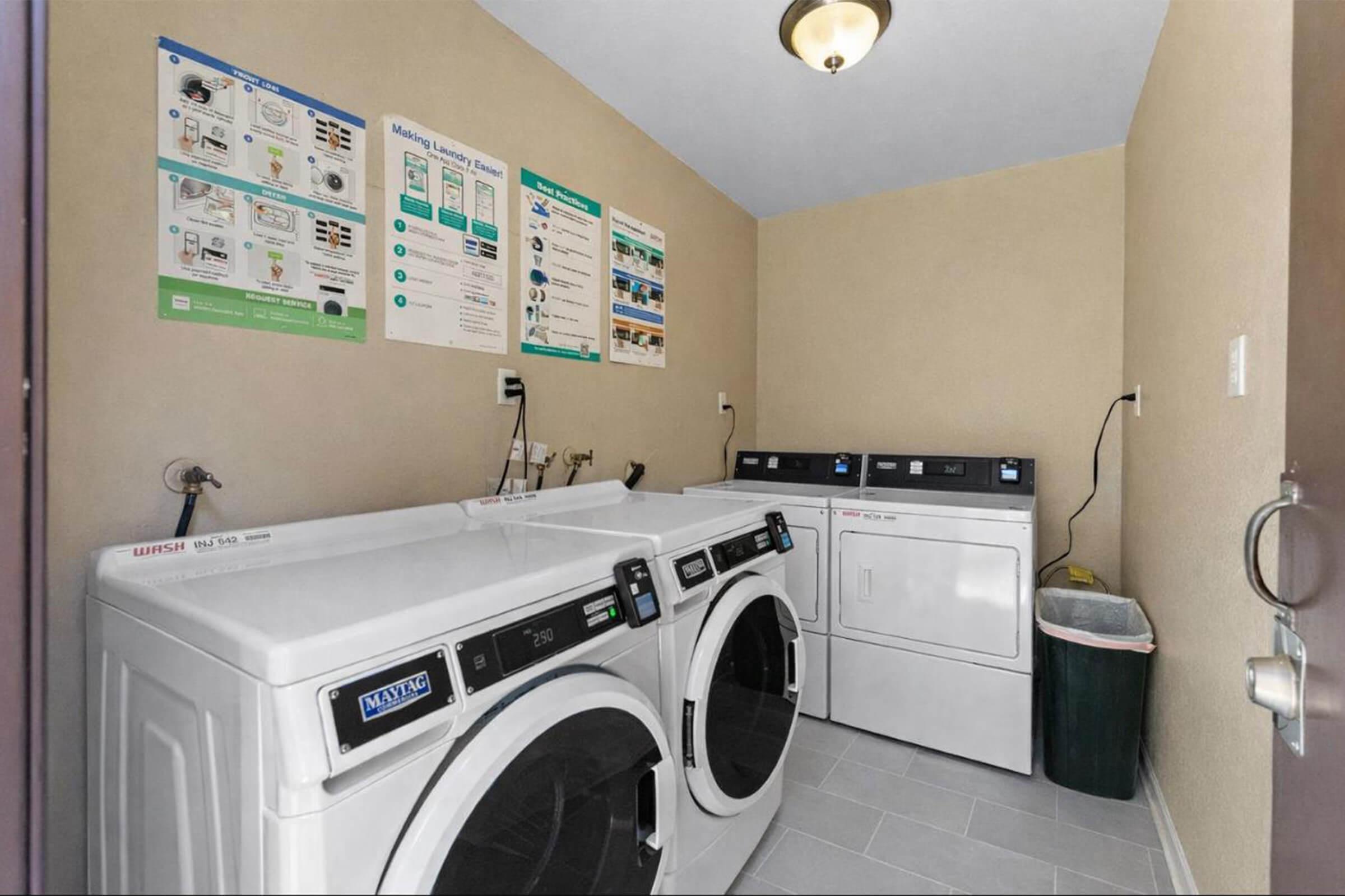 A laundry room featuring three washing machines and a dryer. The machines are stacked, with a wall-mounted light fixture above. Behind the machines, there are informational posters about laundry usage. A green trash bin is positioned in the corner next to the appliances. The walls are painted a neutral color.