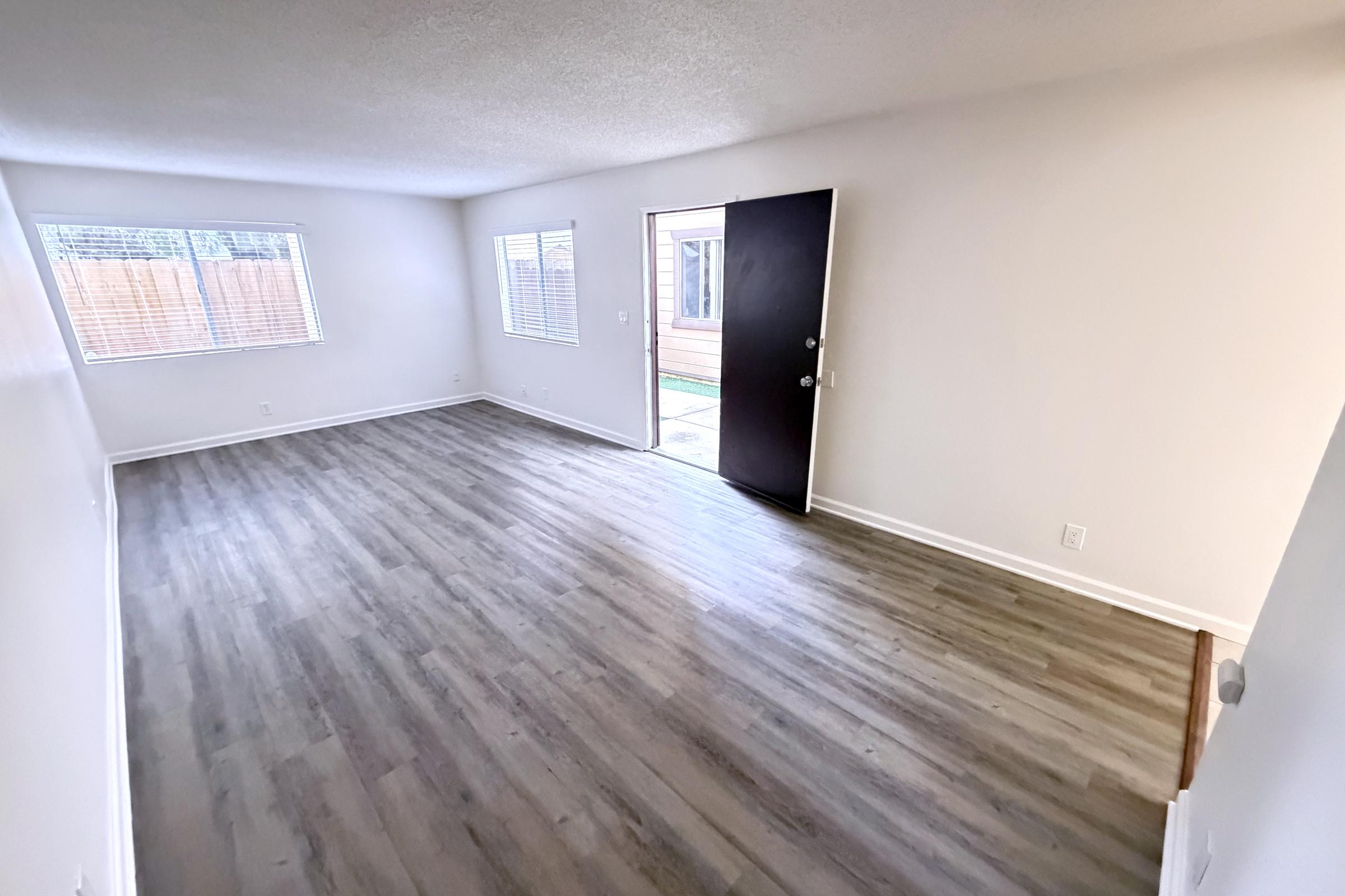 A spacious, empty living room with new laminate flooring, featuring large windows that let in natural light. The room has white walls and a black front door leading outside. The overall atmosphere is bright and airy, ideal for personalizing and decorating.