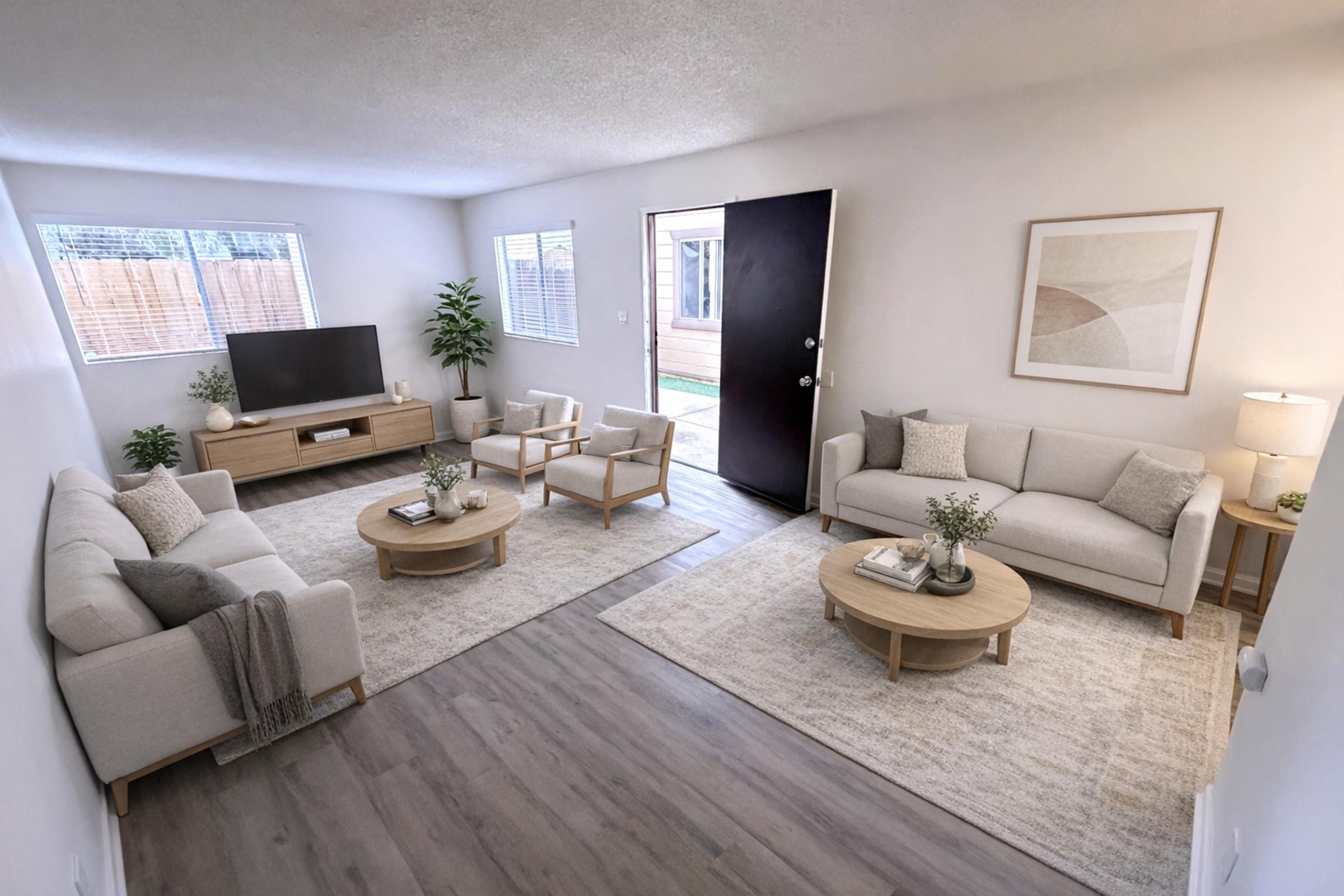 A well-lit, modern living room featuring a cozy seating area with two light-colored sofas and armchairs arranged around two coffee tables. A large TV sits on a wooden console along one wall, with a simple wall art piece above. Natural light streams through the windows, and an inviting door leads outside.