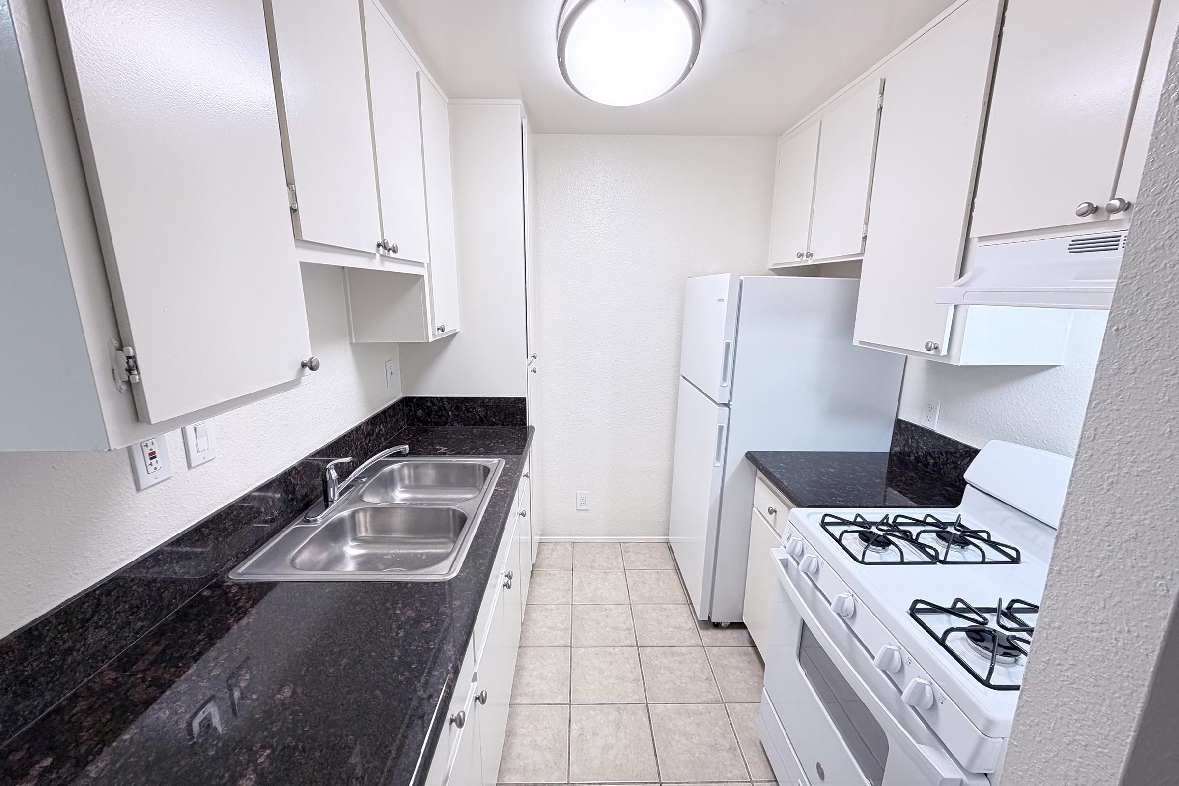 A small, modern kitchen featuring white cabinets, a double sink, a white refrigerator, and a white stove with a gas oven. The countertops are dark granite, and the floor is tiled in light colors. The space is well-lit with a ceiling light.