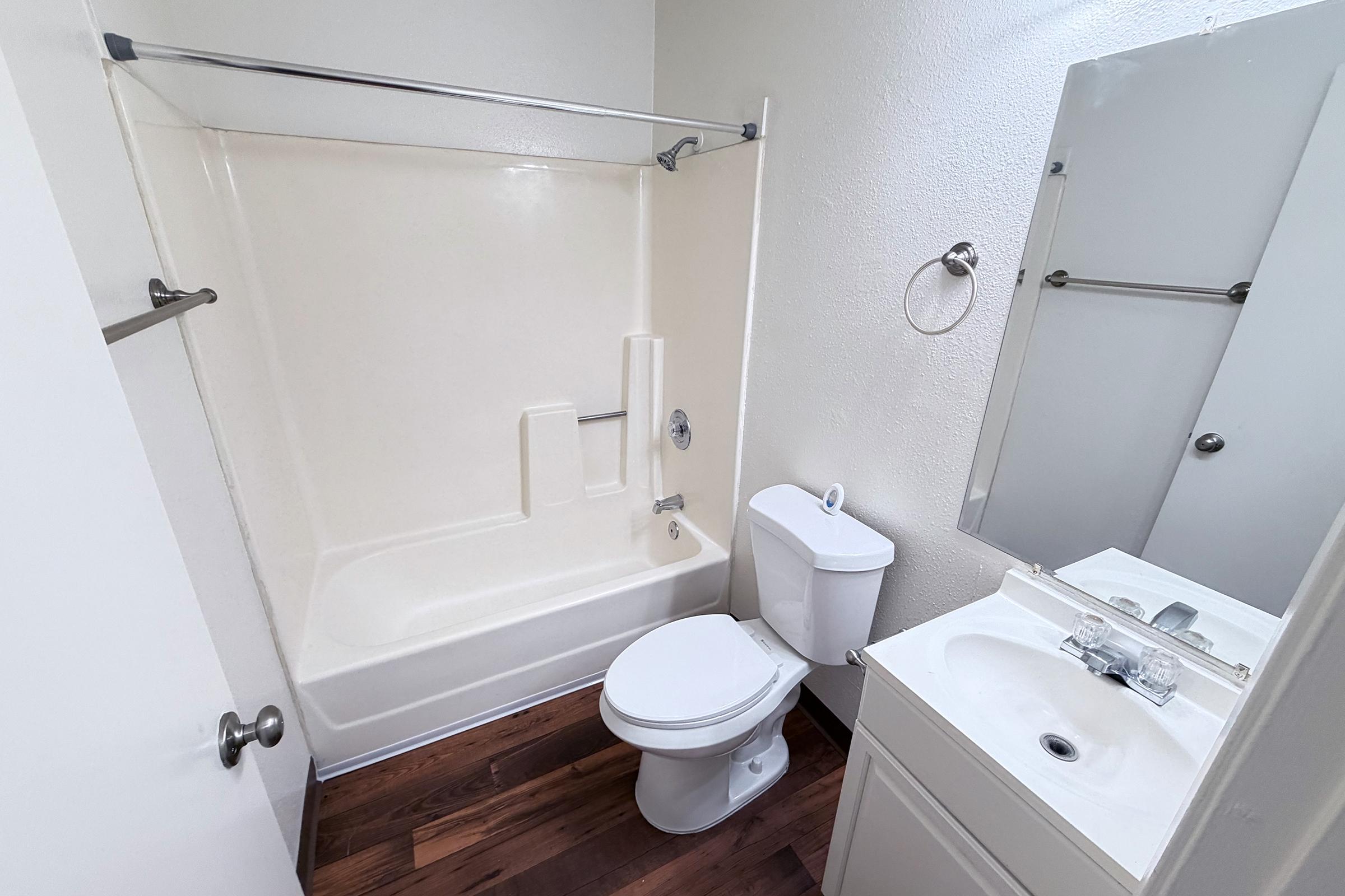 A small bathroom featuring a bathtub with built-in seating, a toilet, and a vanity with a sink. The walls are painted white, and the floor has dark wooden boards. A mirror is mounted above the sink, and a towel rack is visible on the wall. The space is bright and uncluttered.