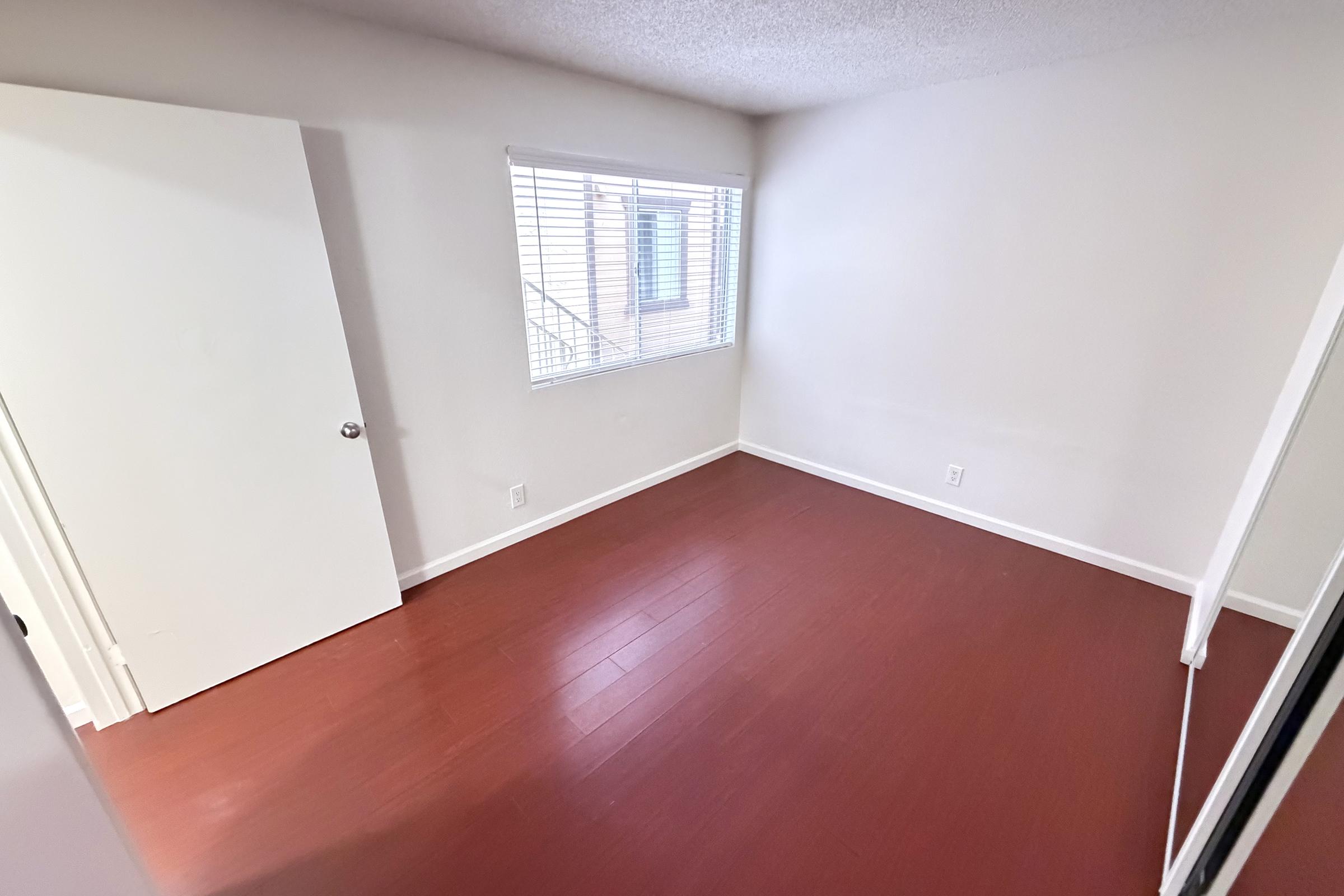 Empty room with red hardwood flooring, a window with blinds allowing natural light in, and a white door partially open. The walls are painted a light neutral color, and there is a mirrored closet door visible.