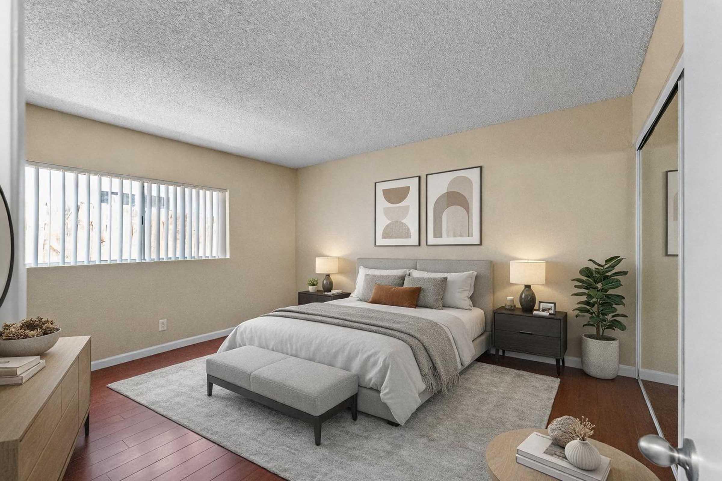 A cozy bedroom featuring a large bed with neutral-colored bedding, a bedside table with lamps, and nature accents. There are two framed artworks on the wall, a window with vertical blinds, and a decorative plant. The room has warm-toned walls and hardwood flooring, creating an inviting atmosphere.