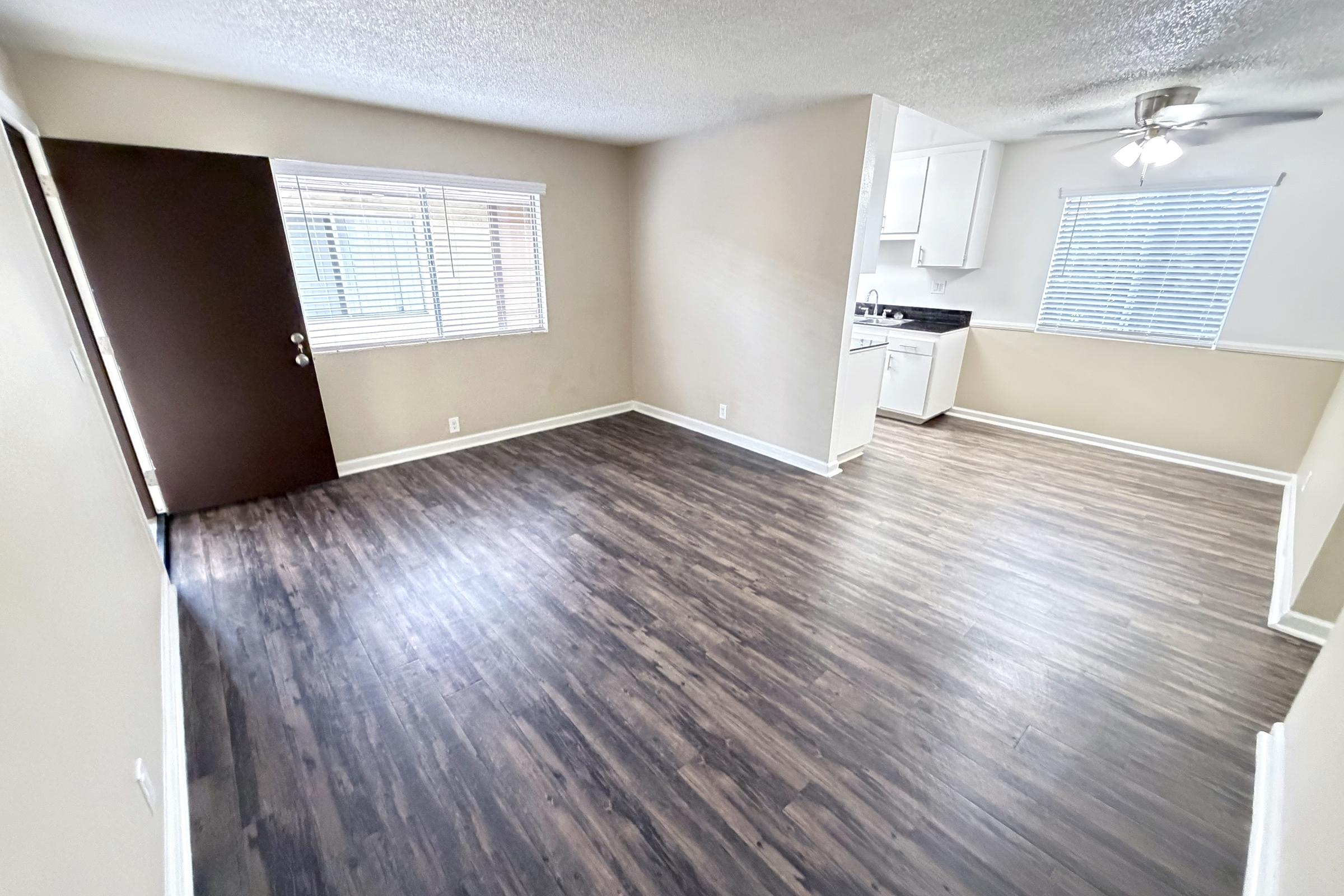 Spacious, well-lit apartment interior featuring laminate flooring, a ceiling fan, and large windows. The room includes an entrance door on the left and a kitchenette visible in the background, with white cabinetry and a black countertop. Walls are painted in a light neutral color.