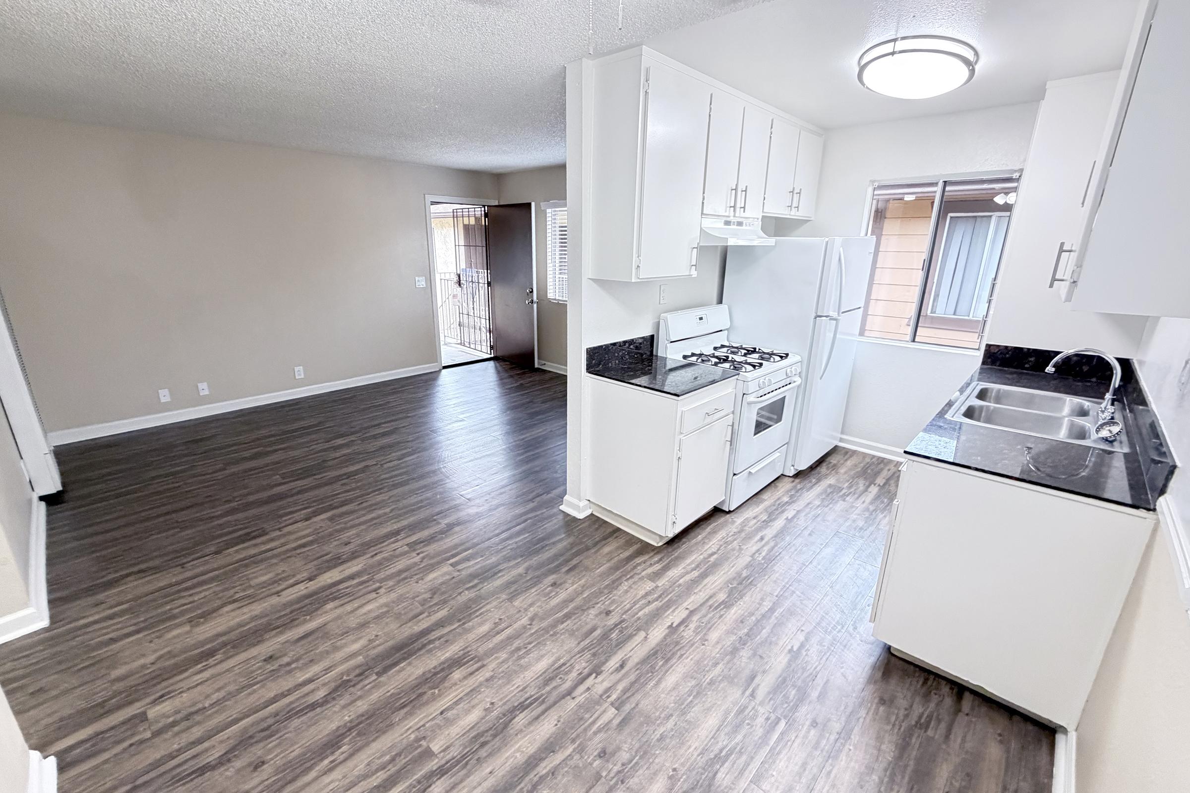 A bright and spacious kitchen with white cabinets and appliances, including a gas stove and sink, adjacent to an open living area with wood-style flooring. Natural light enters through a window, and a door leads to an exterior entryway. The walls are painted in a neutral tone.