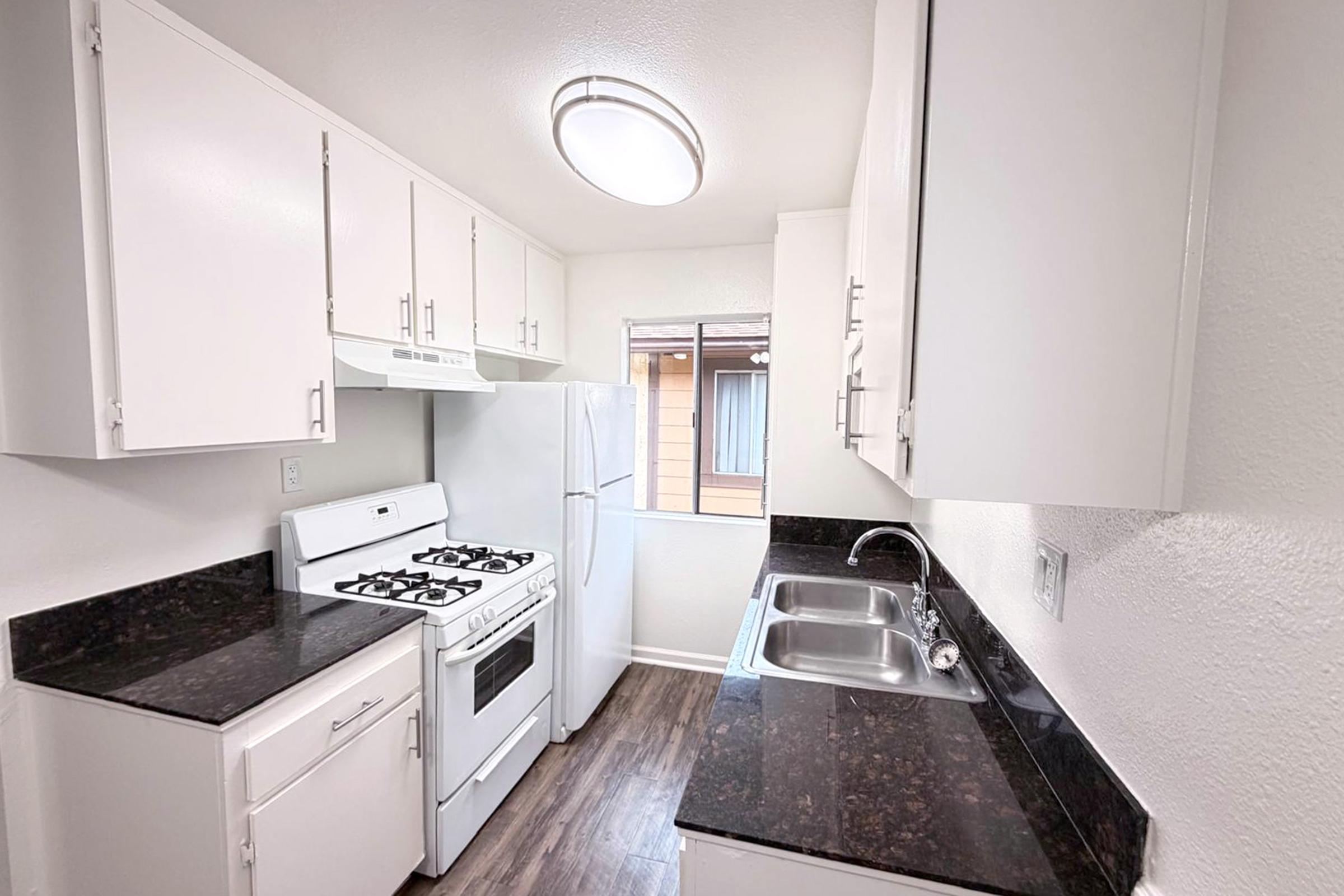 A modern kitchen with white cabinets, a gas stove, a refrigerator, and a stainless steel sink. The countertop has a dark finish, and there is a window providing natural light. The overall design is clean and functional, with a focus on efficient use of space.