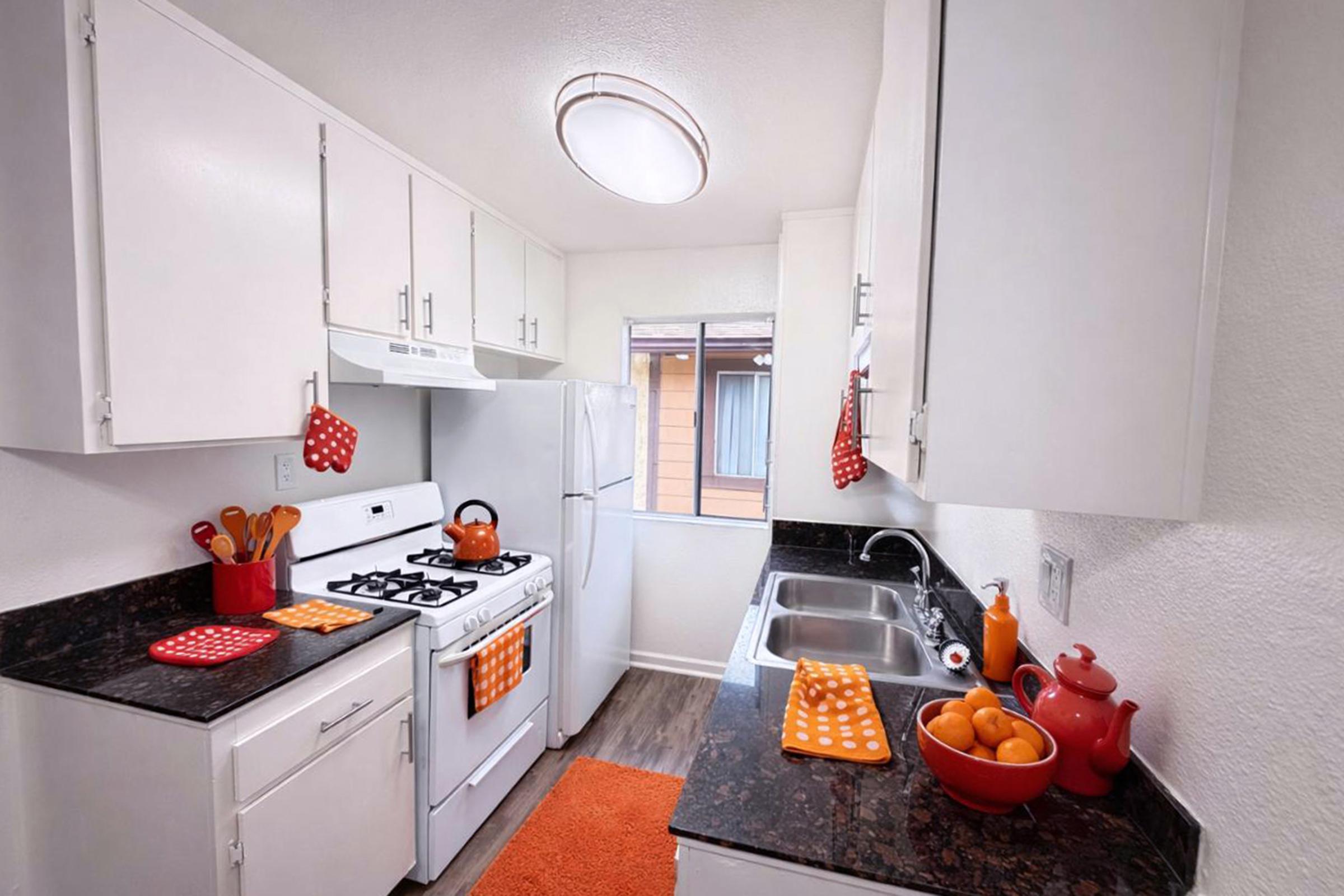A bright, modern kitchen featuring white cabinets, a gas stove, and a stainless steel refrigerator. The countertop is dark granite, adorned with orange accessories, including a teapot and dish towels. A bowl of oranges sits near the sink, and a window lets in natural light, creating a cheerful ambiance.