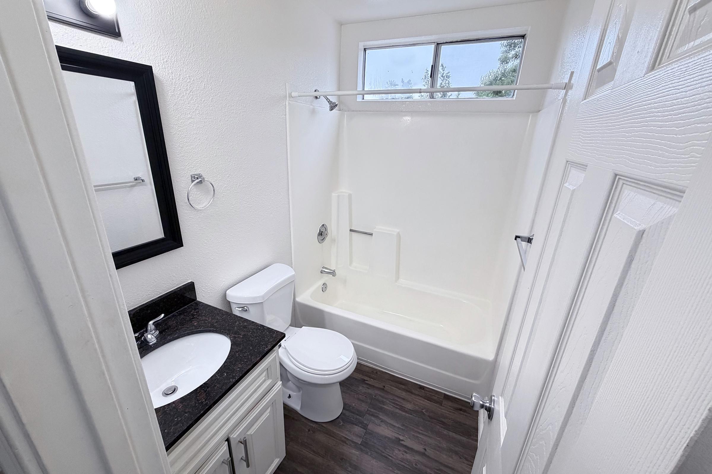 A small, modern bathroom featuring a white tub and shower combination, a black granite countertop with a sink, a toilet, and a large mirror above the sink. The walls are painted white, and there is a window providing natural light. The floor has a dark wood finish.