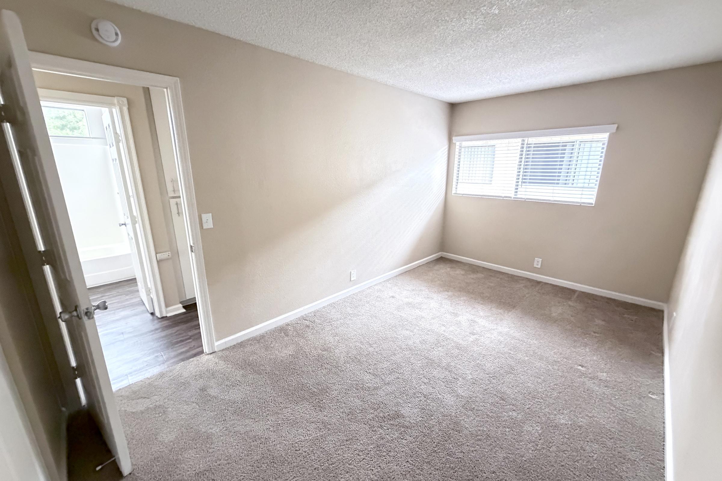 Empty room with light beige walls and carpeted flooring. A window allows natural light to enter, casting shadows inside. A doorway leads to another area, suggesting an adjoining room or bathroom. The space is clean, minimalistic, and ready for furniture or decoration.