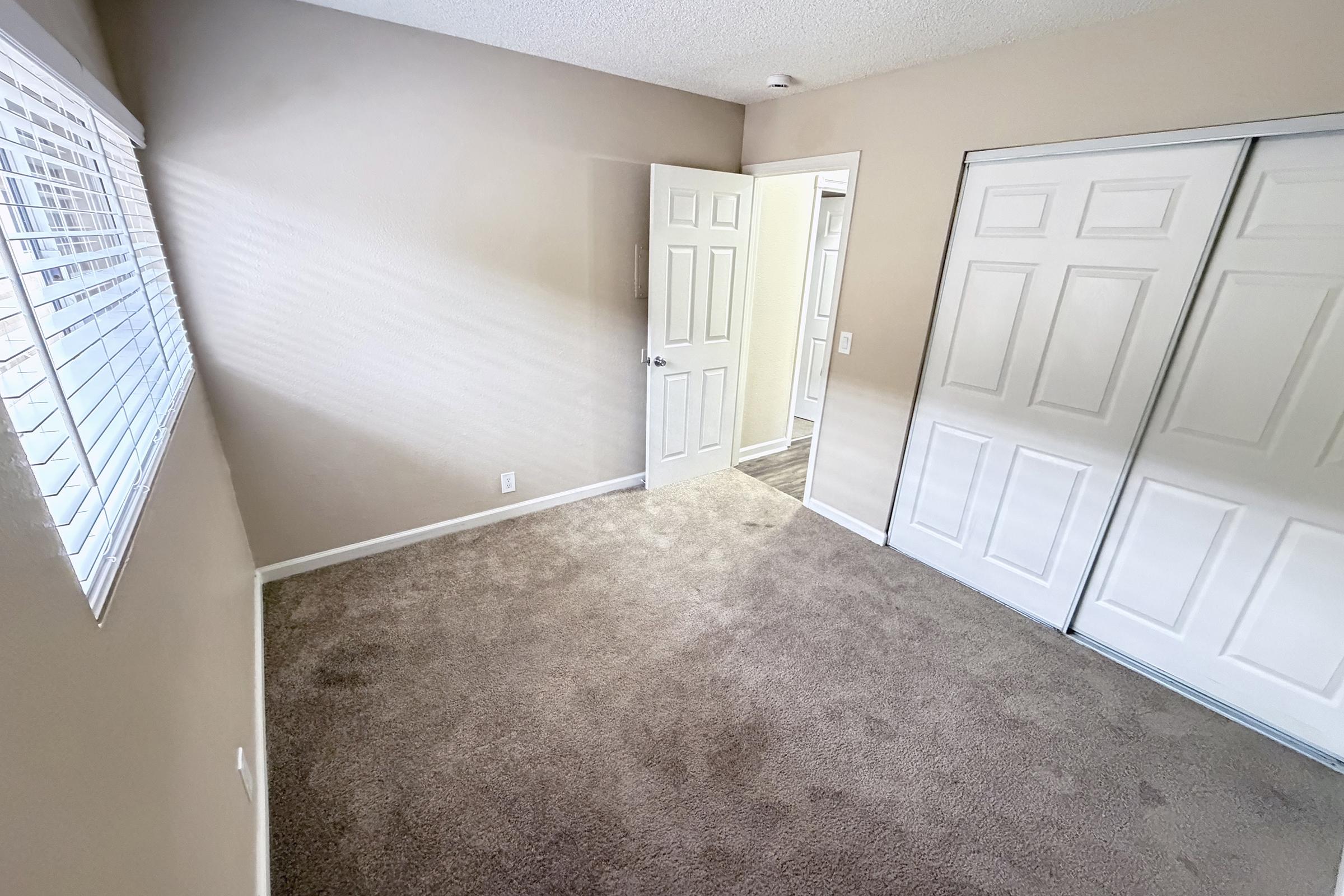 Interior of a small, empty room featuring light beige walls, a carpeted floor, and a window with blinds allowing natural light. A door leads to another room, and there are two closet doors on one wall. The space appears clean and ready for furnishing.