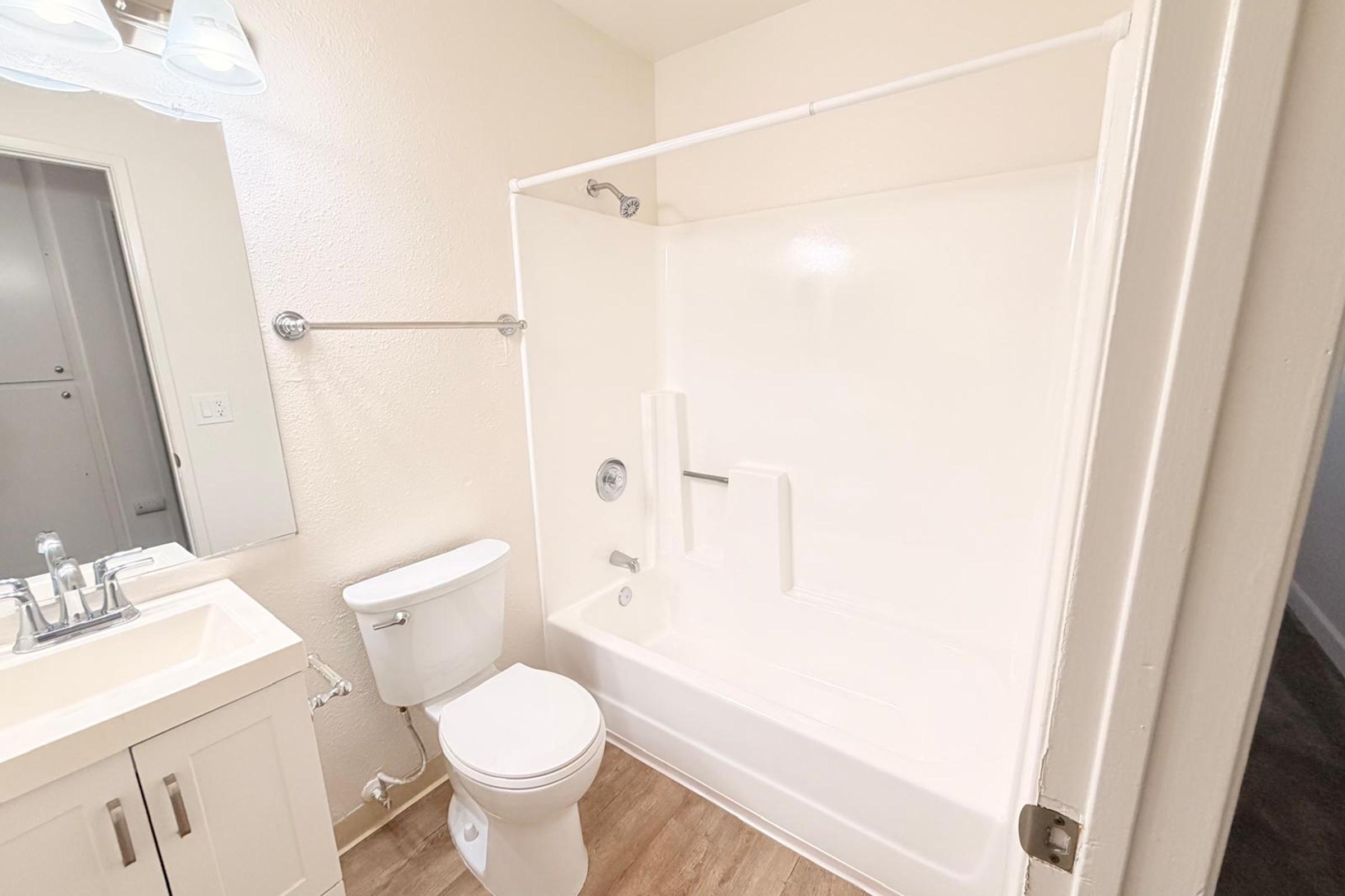 A clean bathroom featuring a white bathtub with a shower, a modern toilet, a sink with a vanity cabinet, and a mirror. The walls are painted light, and the floor is covered with wood-like laminate. A towel bar is visible on the wall near the bathtub.