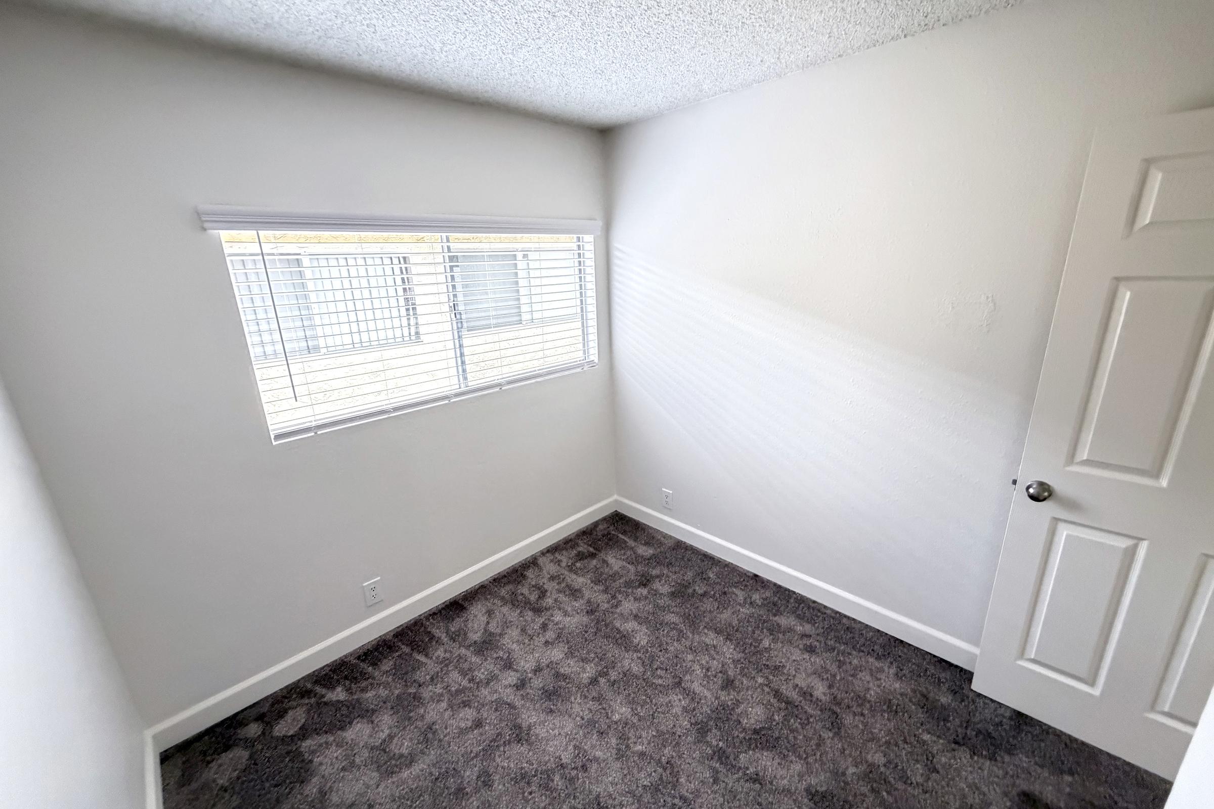 A small, empty room with light gray carpet and white walls. There is a window with closed blinds on the left side, letting in natural light. A white door is visible on the right, and the overall space appears clean and freshly painted.