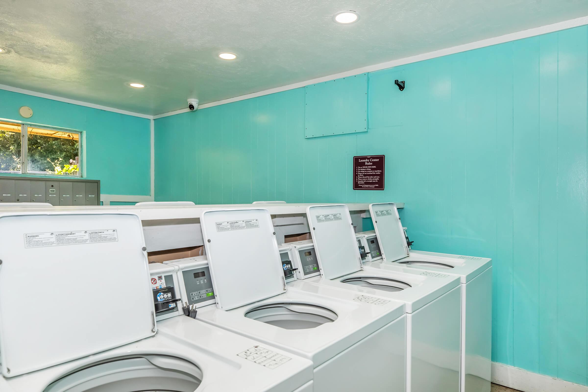 A bright, clean laundry room featuring several white washing machines lined up against turquoise walls. There is a window allowing natural light, and a notice board on the wall. The space is well-lit with ceiling lights, creating a welcoming atmosphere for laundry chores.