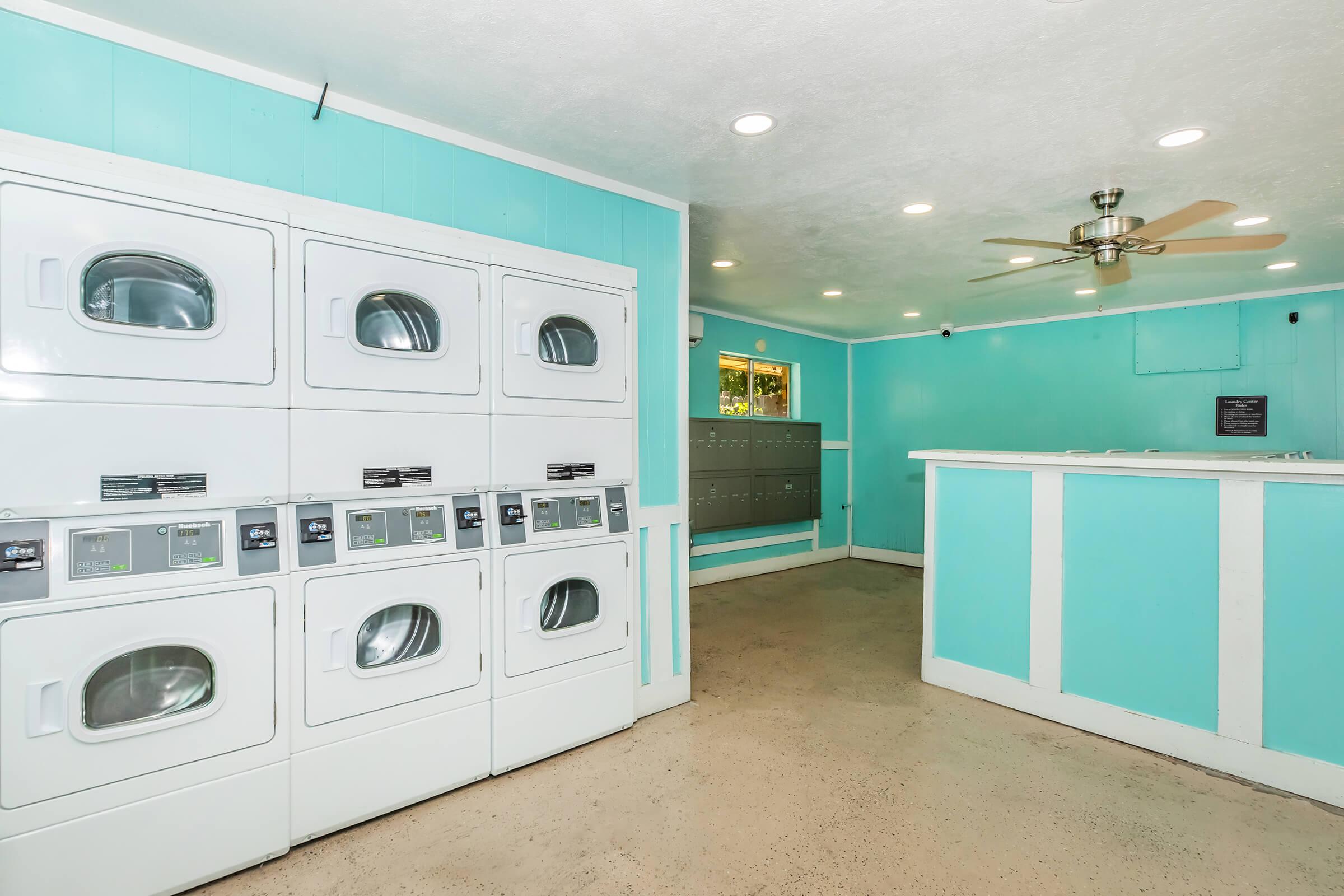 A brightly colored laundry room featuring multiple washing machines and dryers, a mail area with mailboxes, and a service counter. The walls are painted a light turquoise, and there is overhead lighting and a ceiling fan for ventilation. The floor is smooth and neutral-colored.