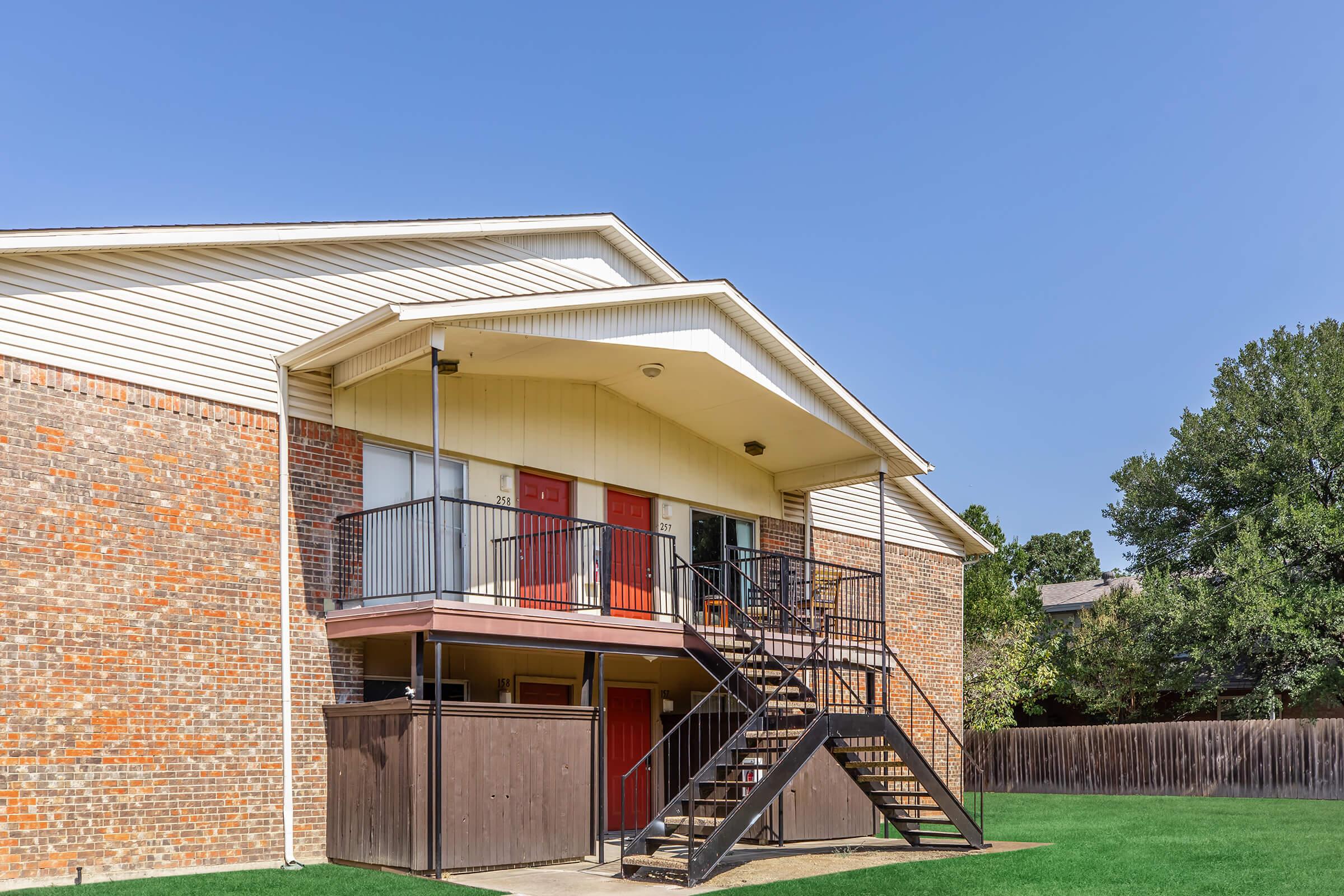 Two-story apartment building with a brick exterior and a sloped roof. The ground floor features a wooden area with stairs leading to the upper level, which has red doors and several large windows. The surrounding grass is well-maintained, and there are trees in the background under a clear blue sky.