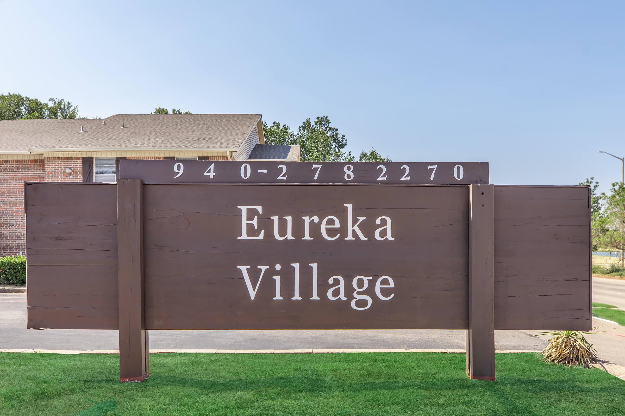 Sign for Eureka Village, featuring large white text on a brown wooden background. The sign includes the phone number "940-278-2270" and is located near a grassy area, with a clear blue sky in the background.