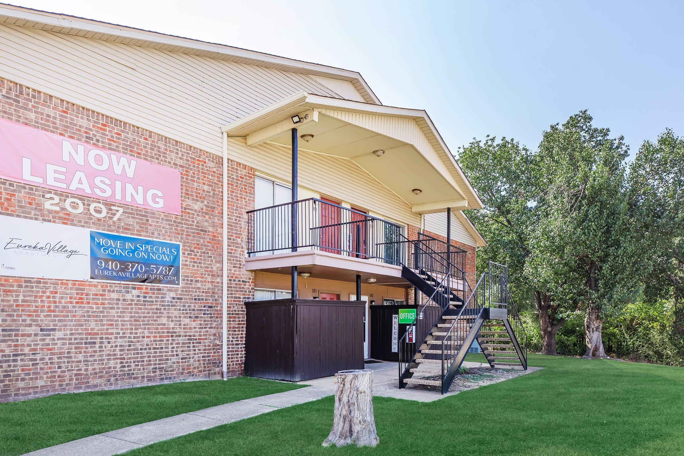 Exterior view of a leasing office for an apartment complex. The building features a brick facade, a balcony with black railings, and stairs leading to the entrance. A large "Now Leasing" sign is displayed prominently, and there is a well-maintained lawn in front. Trees are seen in the background under a clear sky.
