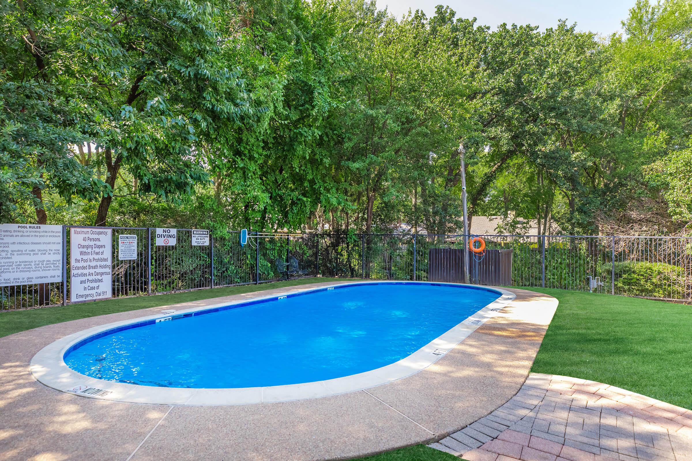 A clear blue swimming pool surrounded by green trees and grass. The area is enclosed by a fence, with safety signs visible nearby. The pool features a ladder and a lifebuoy on the fence. Sunlight is shining, creating a tranquil atmosphere.