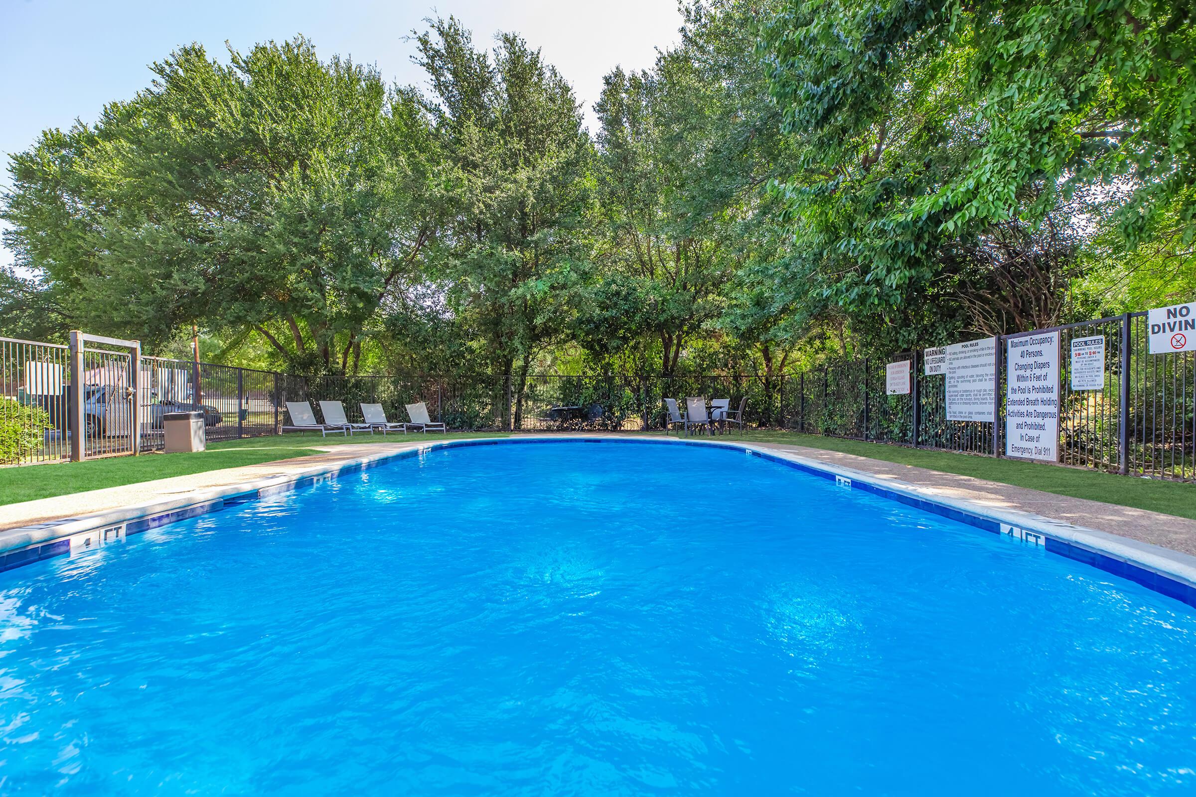 A clear blue swimming pool surrounded by lush green trees. There are lounge chairs on one side of the pool and several informational signs posted around. The atmosphere is tranquil and inviting, perfect for relaxation and enjoyment.