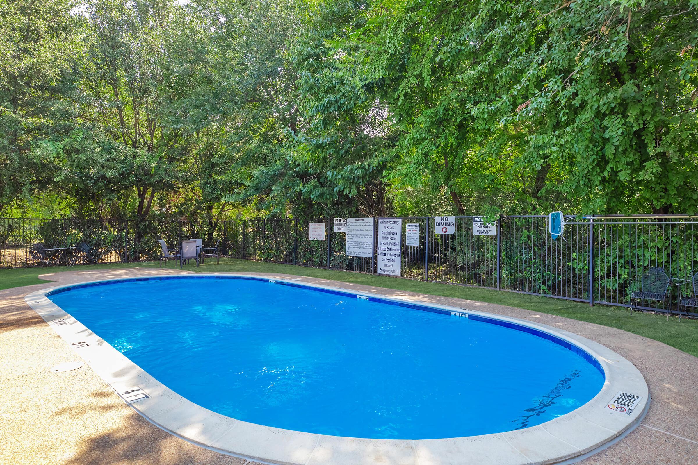A clear blue swimming pool surrounded by lush greenery. The pool has a curved shape and is bordered by a light-colored concrete deck. There are several signs posted around the pool area, and a single chair is visible nearby. The scene is peaceful, indicating a sunny day perfect for swimming or relaxation.