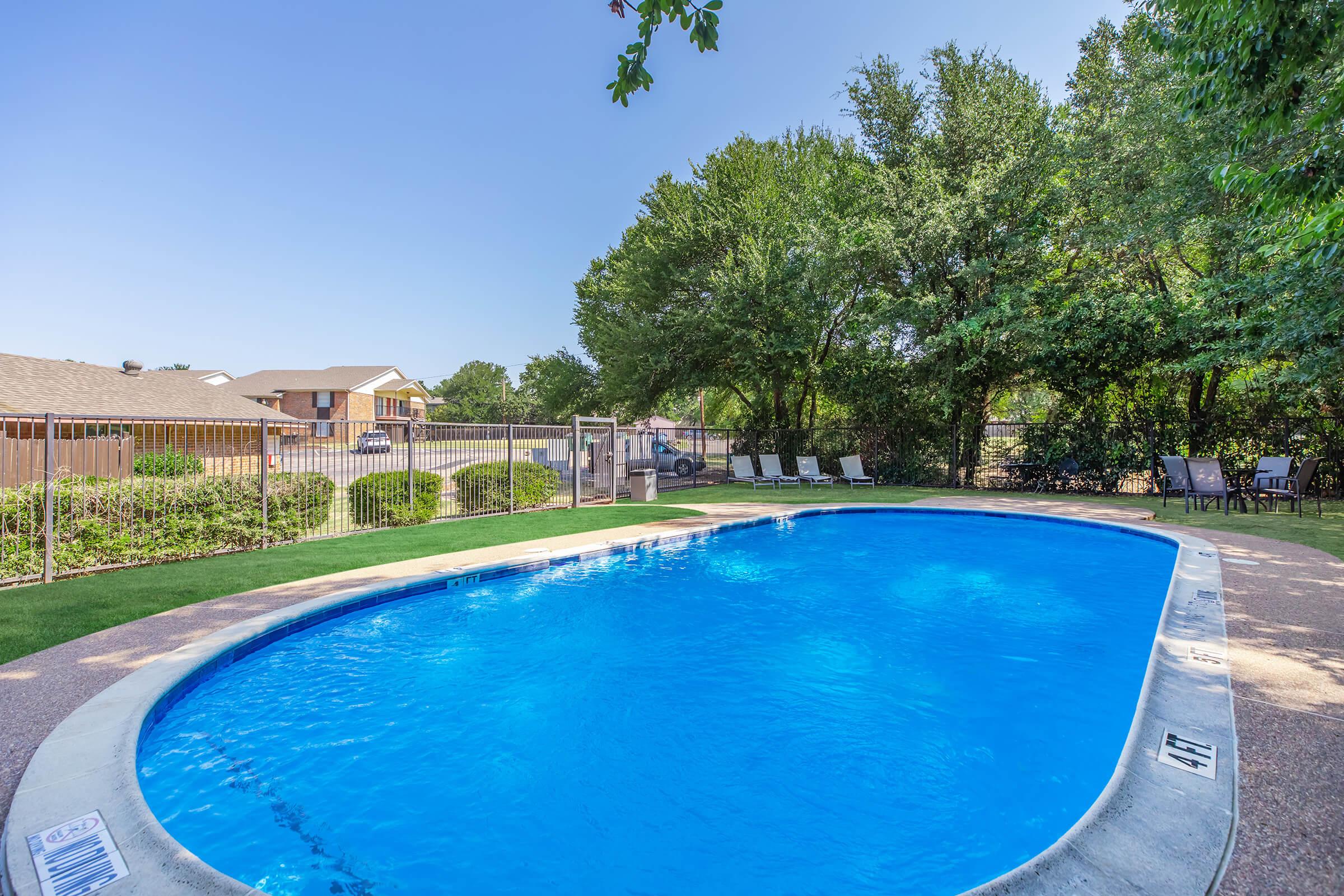 A clear blue swimming pool surrounded by lush green trees and a fenced area. Lounge chairs are set up nearby on the grass, creating a relaxing outdoor space. In the background, residential buildings are visible. The sky is clear and sunny, adding to the inviting atmosphere.
