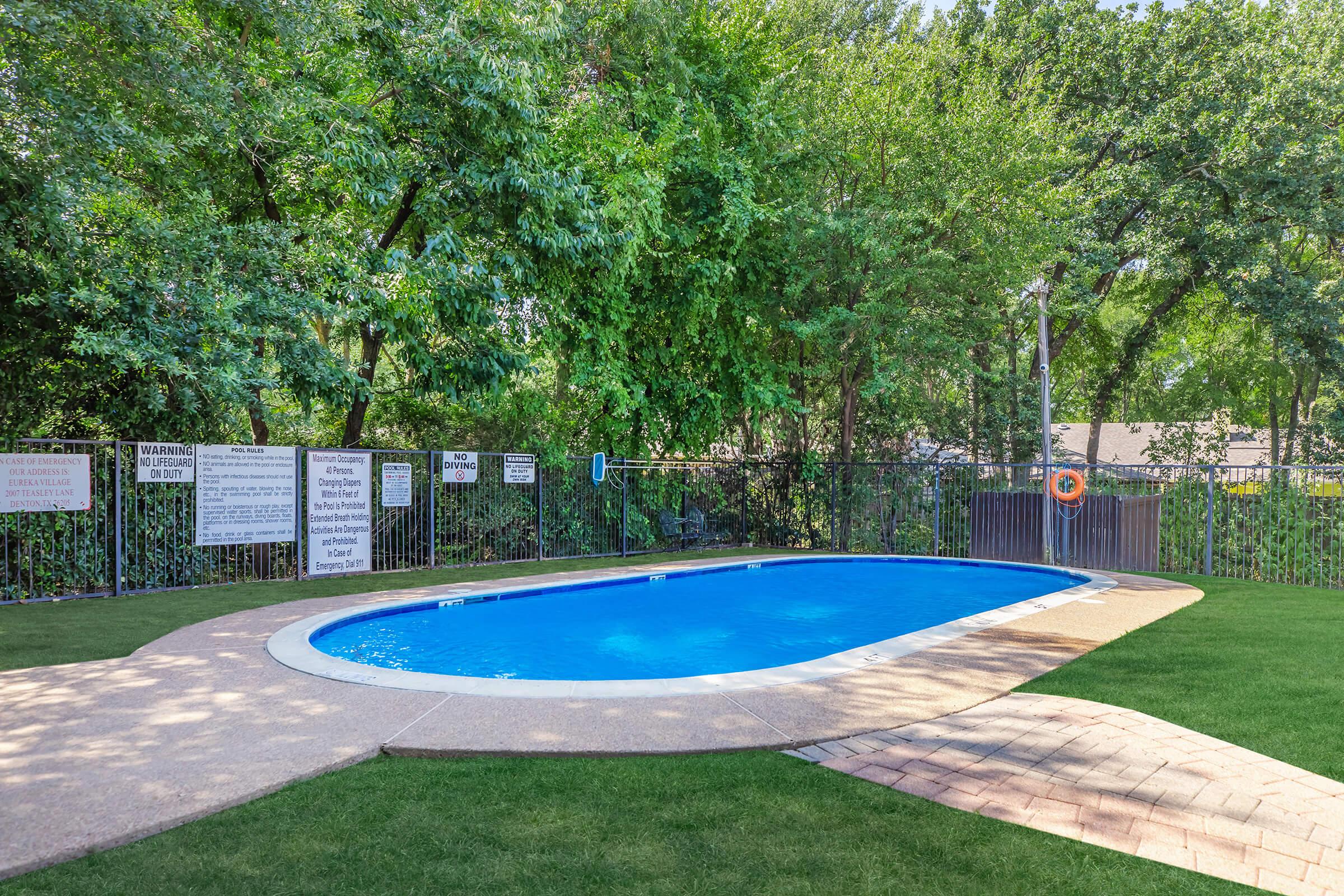 A small in-ground swimming pool surrounded by lush greenery and a wooden fence. The pool has clear blue water, a light-colored surrounding deck, and signs posted around the perimeter, including safety warnings. The area is well-maintained with neatly trimmed grass.