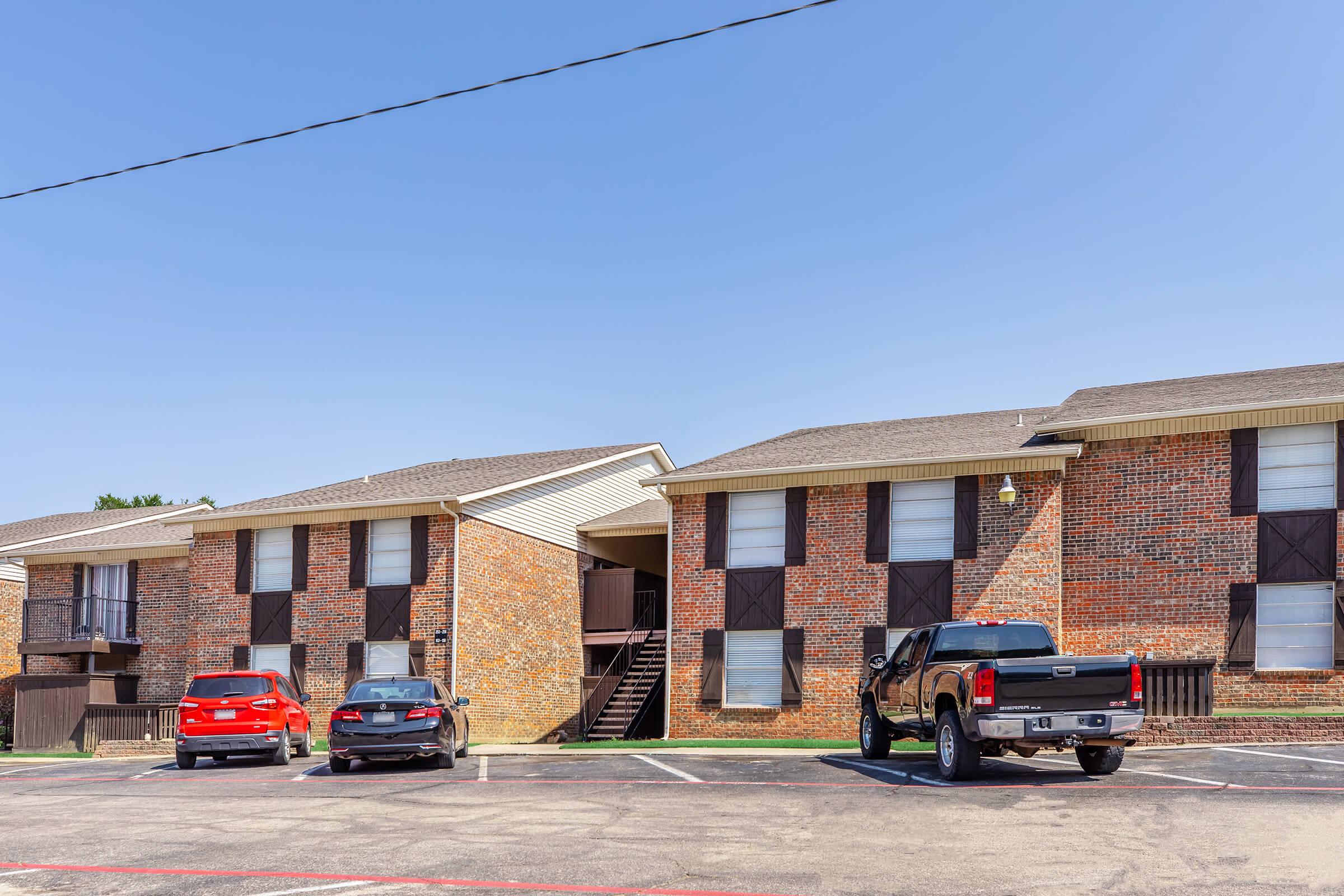 A brick multi-unit apartment building with two levels, featuring several parked cars in the foreground, including a red car and a black truck. The sky is clear and blue, and the building has brown shutters on the windows. There's a staircase leading to the upper level.
