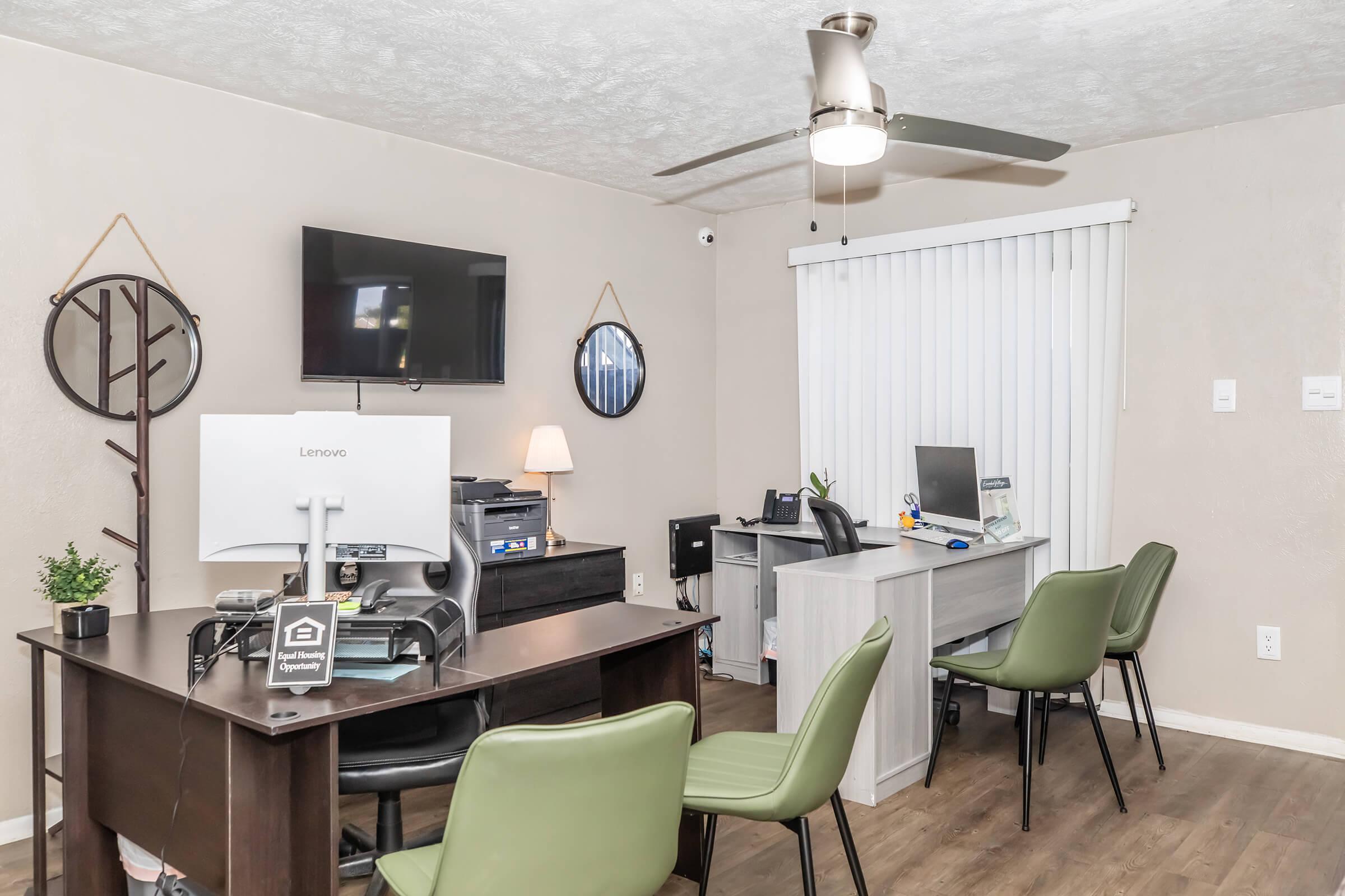 An office space featuring a brown desk with a computer, a modern chair, two green chairs, and a gray desk. The room has a wall-mounted TV, a ceiling fan, and large window blinds. Decorative items include a plant and round mirrors on the walls, creating a professional and inviting atmosphere.