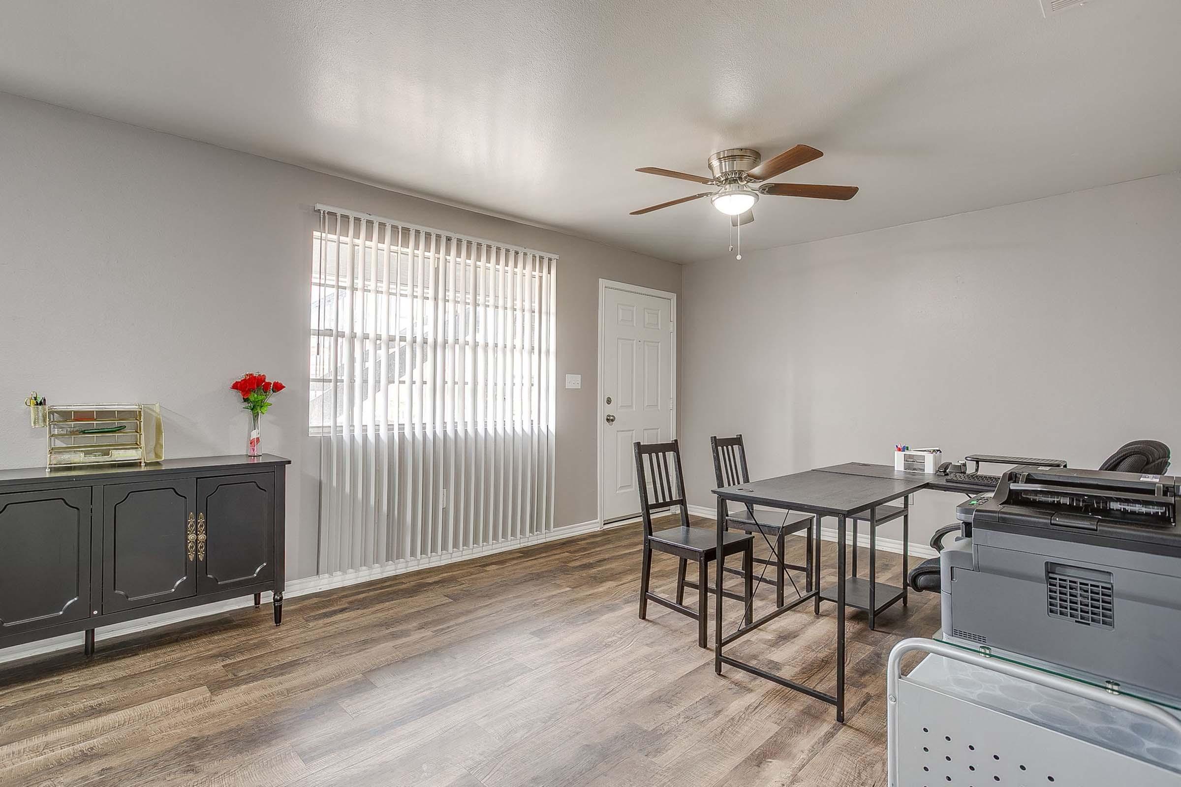 A simple, modern room featuring a table with four chairs, a black cabinet, and a small decorative plant. A ceiling fan is installed, and there are blinds on the window allowing natural light to enter. A printer is visible on the side, contributing to a functional workspace environment.