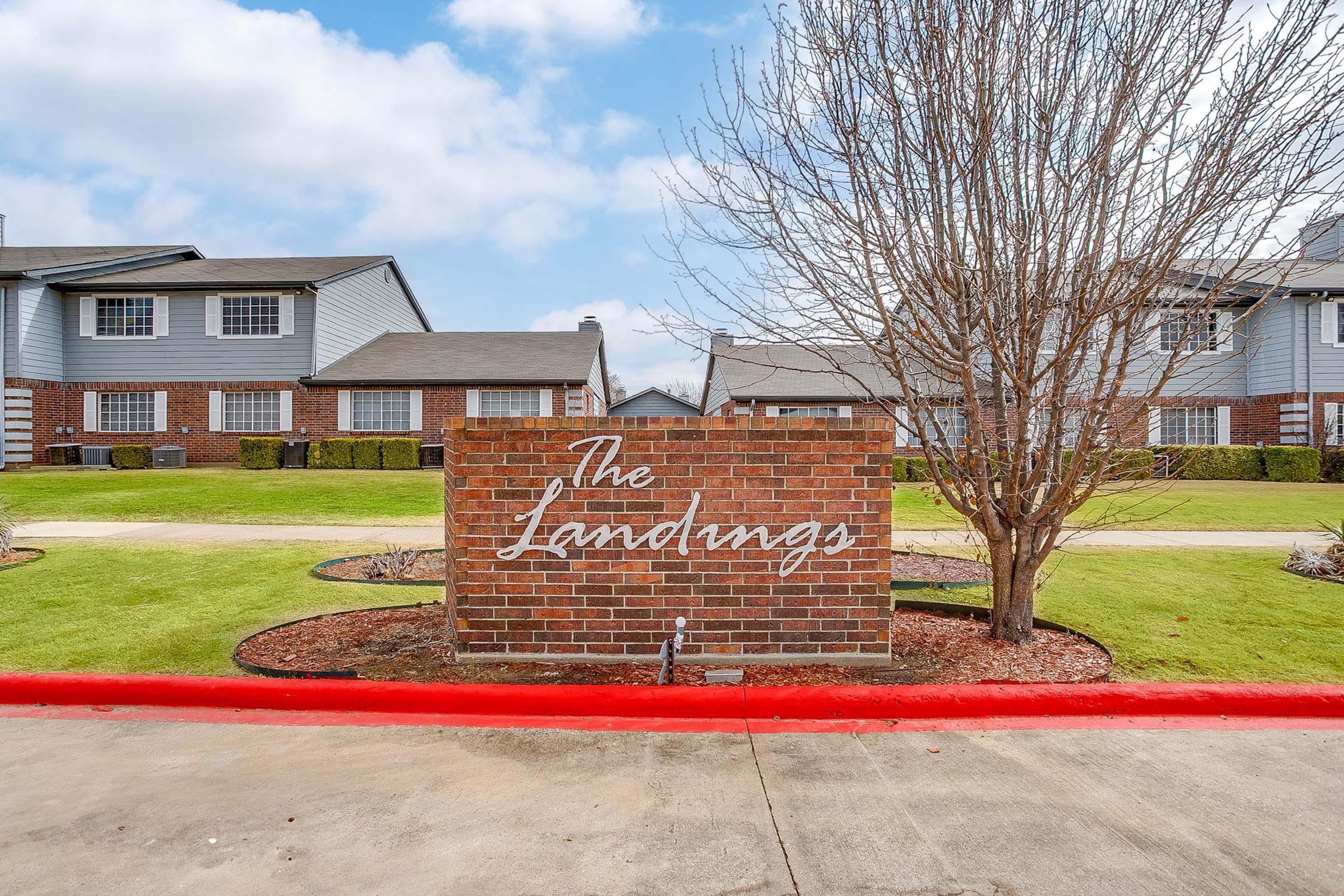 A welcome sign for "The Landings," featuring a brick structure with the name elegantly displayed. The background includes several residential buildings, neatly trimmed shrubbery, and a clear blue sky. The area has a well-maintained lawn with a red curb.