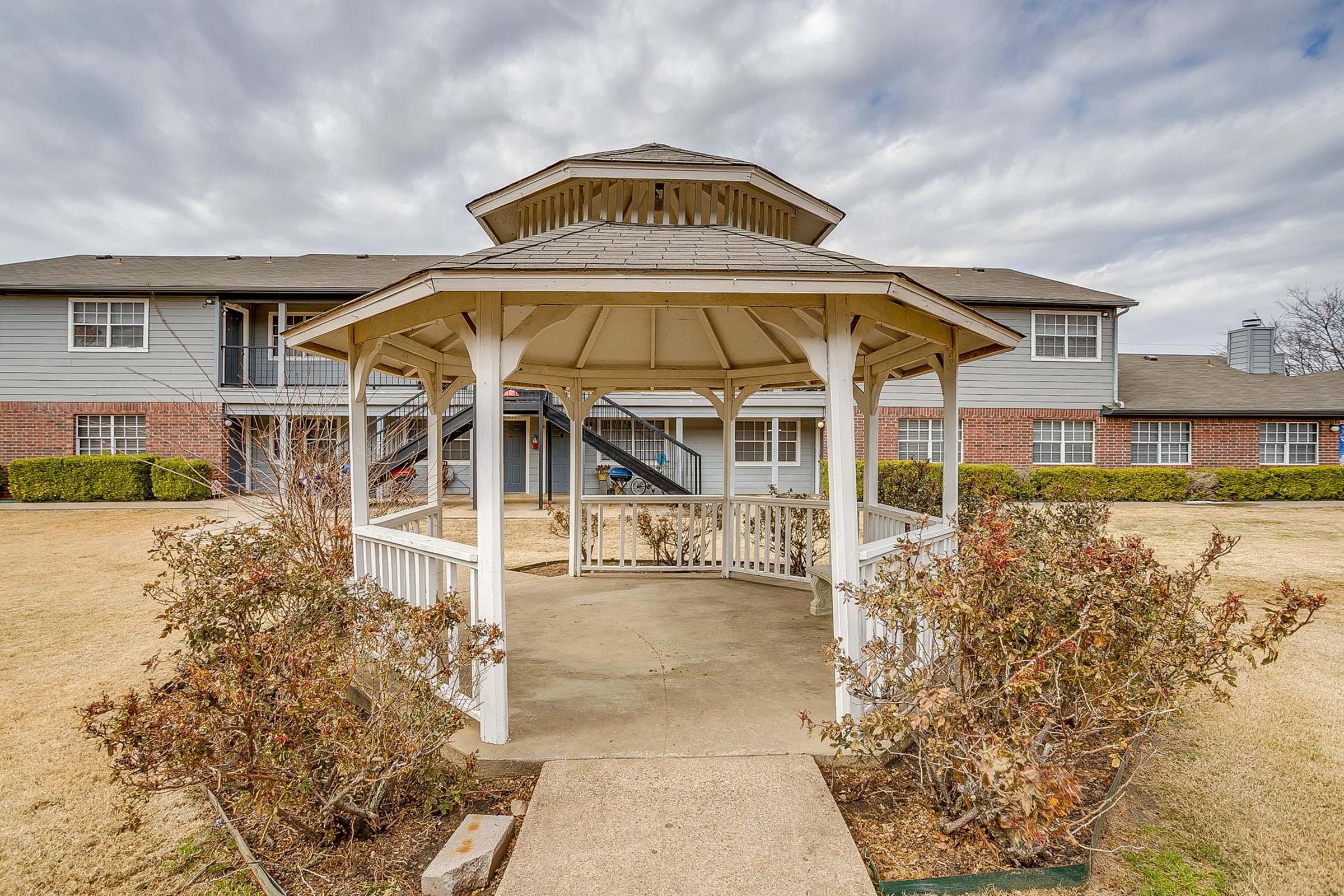 A white gazebo with a wooden structure and a shingled roof is situated in the foreground, surrounded by shrubs and grass. In the background, there are two-story apartment buildings and a cloudy sky, creating a tranquil outdoor setting.