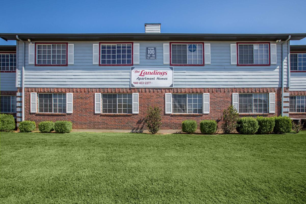 Exterior view of The Landings Apartment Homes, featuring a two-story building with a green lawn in front. The building has large windows, a brick facade, and a prominent sign displaying the name and contact number for the apartments. Clear blue sky above.
