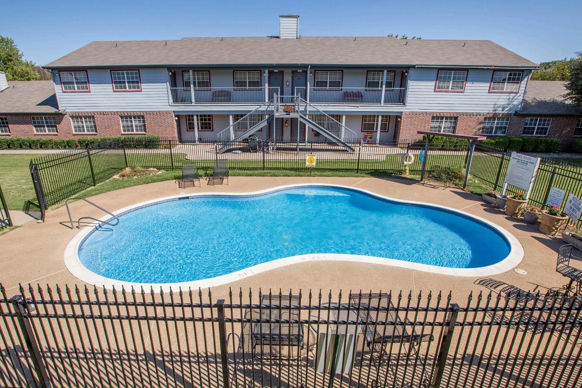 A view of a pool in an outdoor area surrounded by a black fence. The pool is shaped like a freeform figure and features clear blue water. In the background, there is a two-story building with multiple apartments visible, and well-maintained grass and landscaping around the pool area.