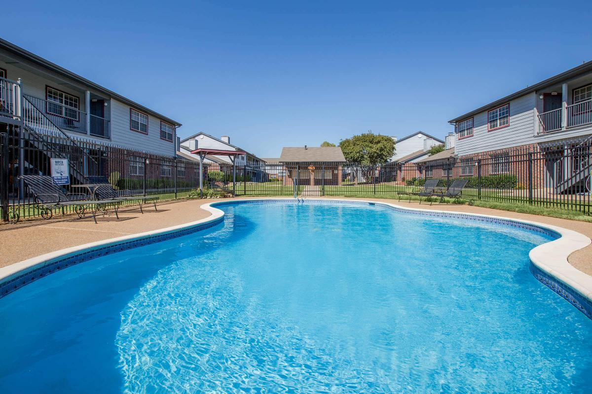 A clear blue swimming pool surrounded by a fenced area, with lounge chairs positioned nearby. The setting features two-story apartment buildings in the background, set against a clear blue sky, with green grass and landscaping around the pool area.