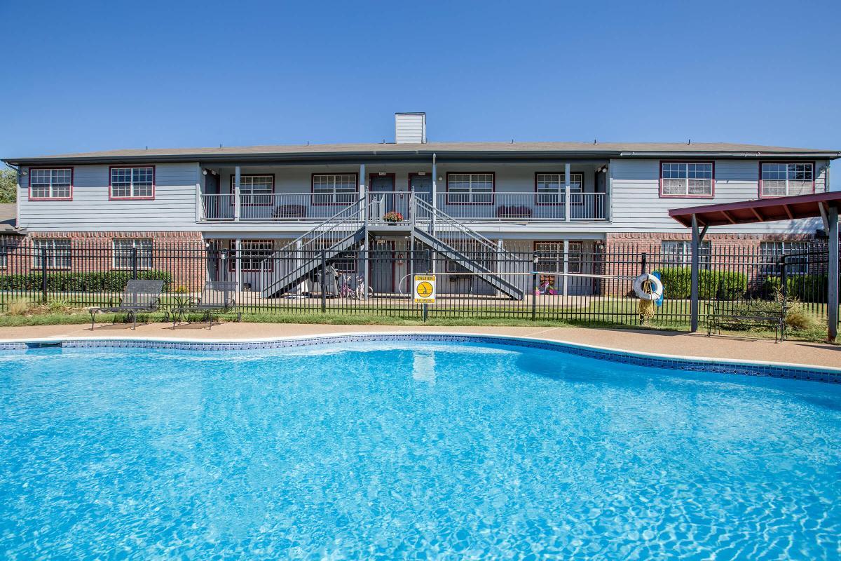 A clear view of an apartment building with multiple floors, framed by a blue swimming pool in the foreground. The building has a light blue exterior and balconies, with some decorative elements. There is a fence around the pool area, and the sky is bright and sunny.