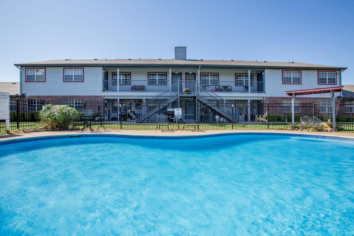 A view of a multi-unit residential building with stairs leading up to apartments, situated behind a swimming pool. The pool is clear and inviting, surrounded by a fence and green landscaping, under a clear blue sky.