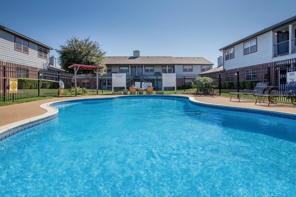 A bright blue swimming pool surrounded by green grass and lounge chairs. In the background, there are two-story apartment buildings with a clear blue sky overhead. A shaded area with a red canopy is visible near the pool, creating a relaxing atmosphere.