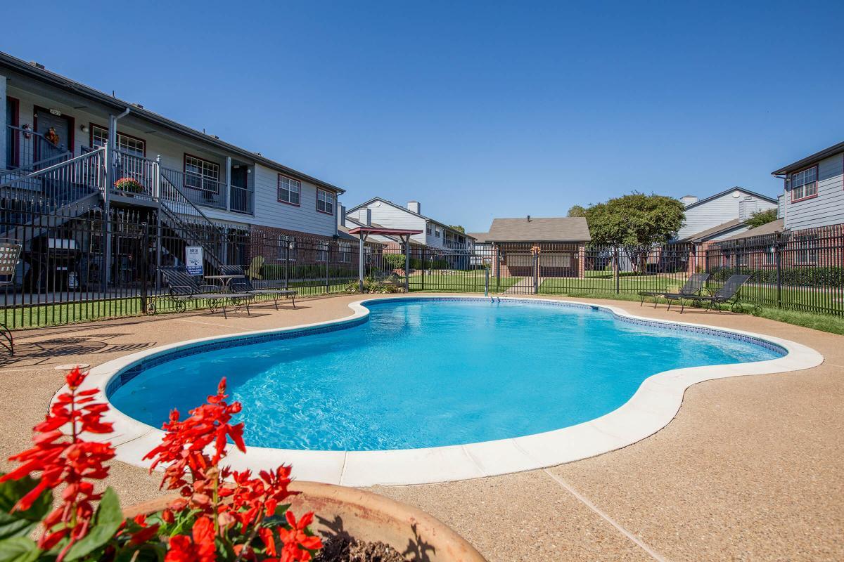 A clear blue swimming pool surrounded by a fenced area, with lounge chairs positioned around it. In the foreground, there are vibrant red flowers in a pot. The background features multi-story residential buildings and manicured green lawns under a clear blue sky.