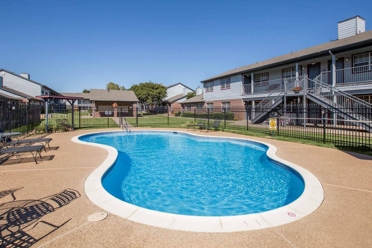 A clear blue swimming pool shaped like a kidney bean, surrounded by a sandy-colored patio. In the background, there are two-story apartment buildings with balconies and a well-maintained grassy area. The sky is bright and clear, indicating a sunny day.