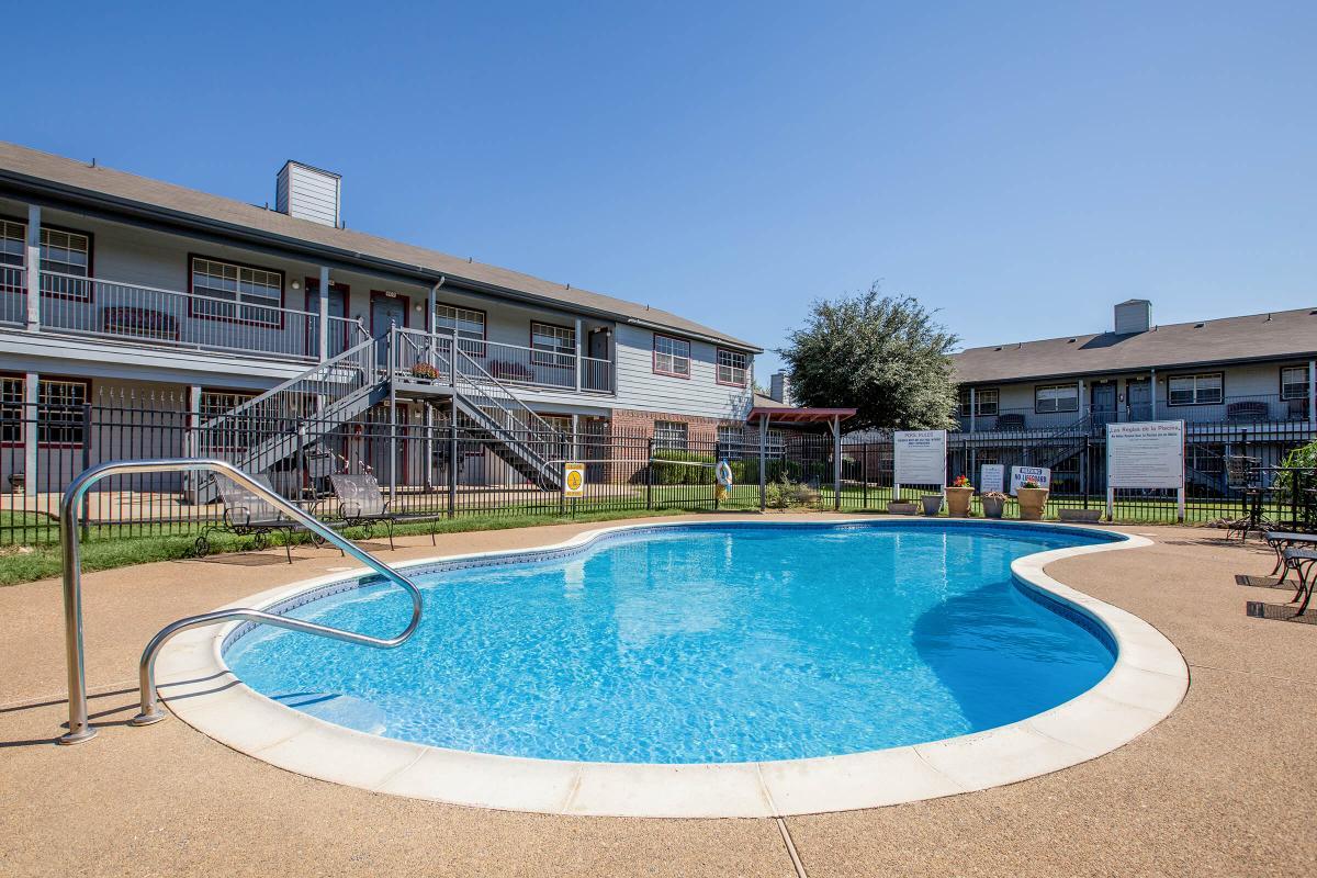 A swimming pool in a residential complex, featuring a curved shape and surrounded by a concrete deck. In the background, a two-story building with balconies is visible, along with lounge chairs and a fenced area. Clear blue skies add to the ambiance of the outdoor space.