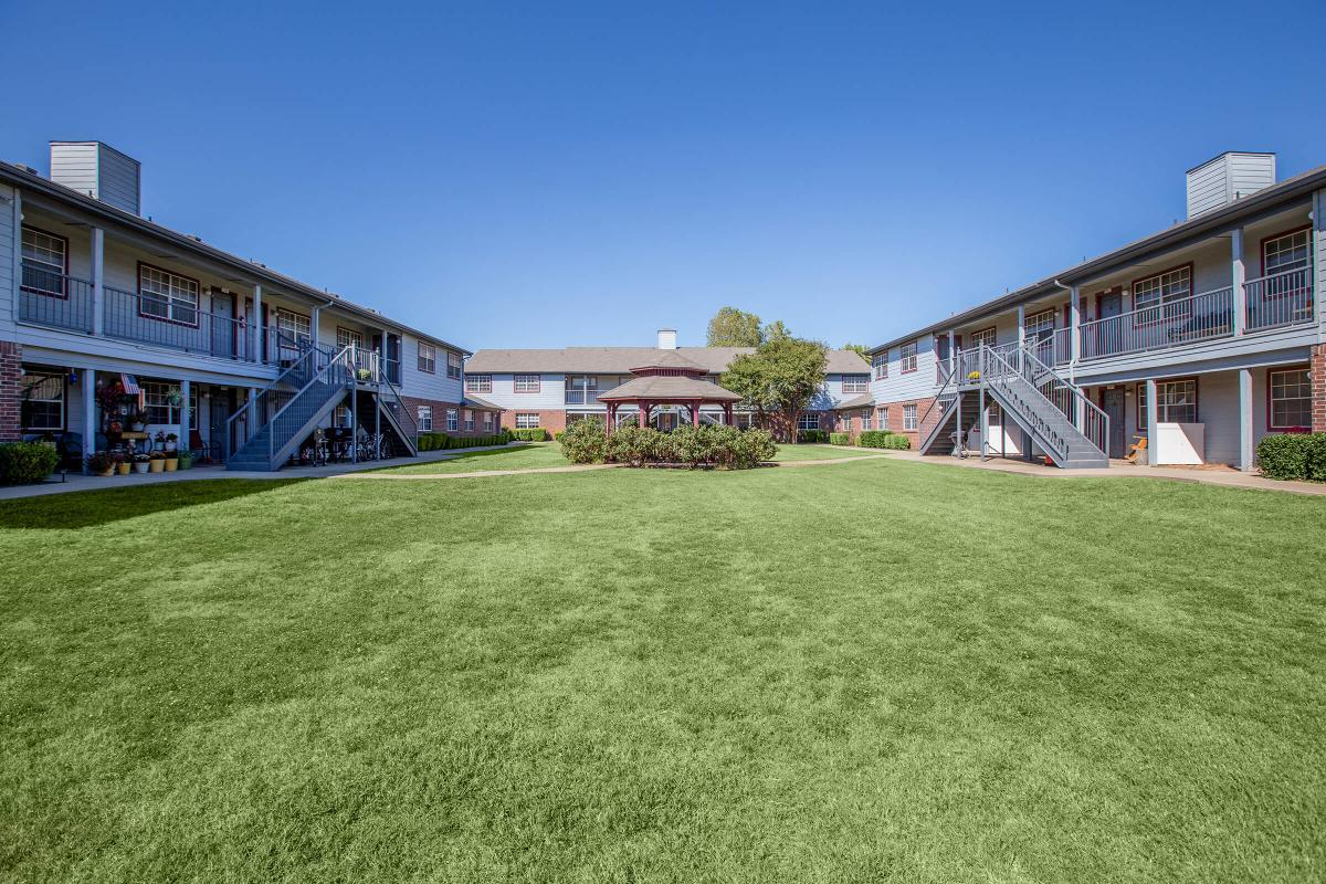 View of a well-maintained courtyard in an apartment complex, featuring lush green grass and several buildings with balconies. In the center, there is a gazebo surrounded by shrubs, providing a communal space. The sky is clear and blue, creating a bright and inviting atmosphere.