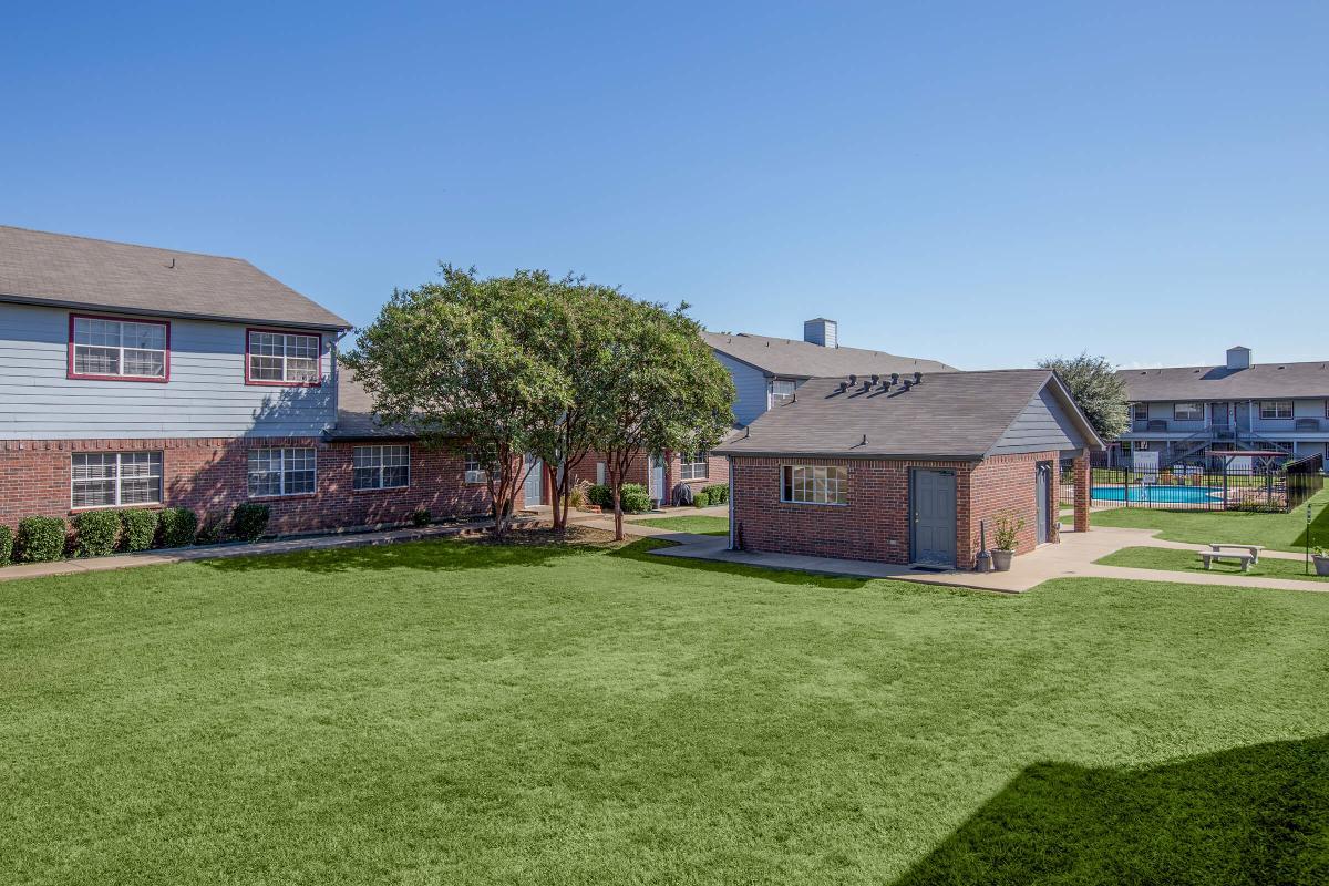 View of a sunny outdoor area with well-maintained grass, surrounded by residential buildings. In the background, a swimming pool is visible, along with a fence. Several trees provide shade, and there are benches for seating. The overall setting conveys a relaxed, community atmosphere.